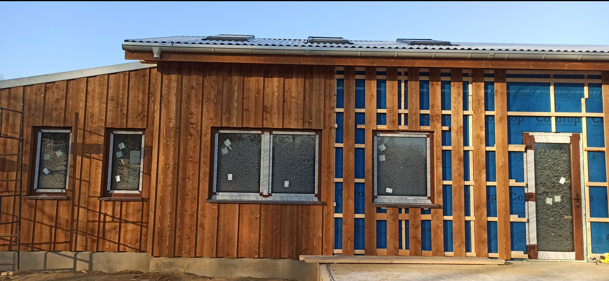 Budowa domu szkieletowego z drewnianą elewacją w trakcie montażu. Widoczne okna, drzwi i fragmenty konstrukcji. Część elewacji bez desek, ukazująca niebieską membranę.