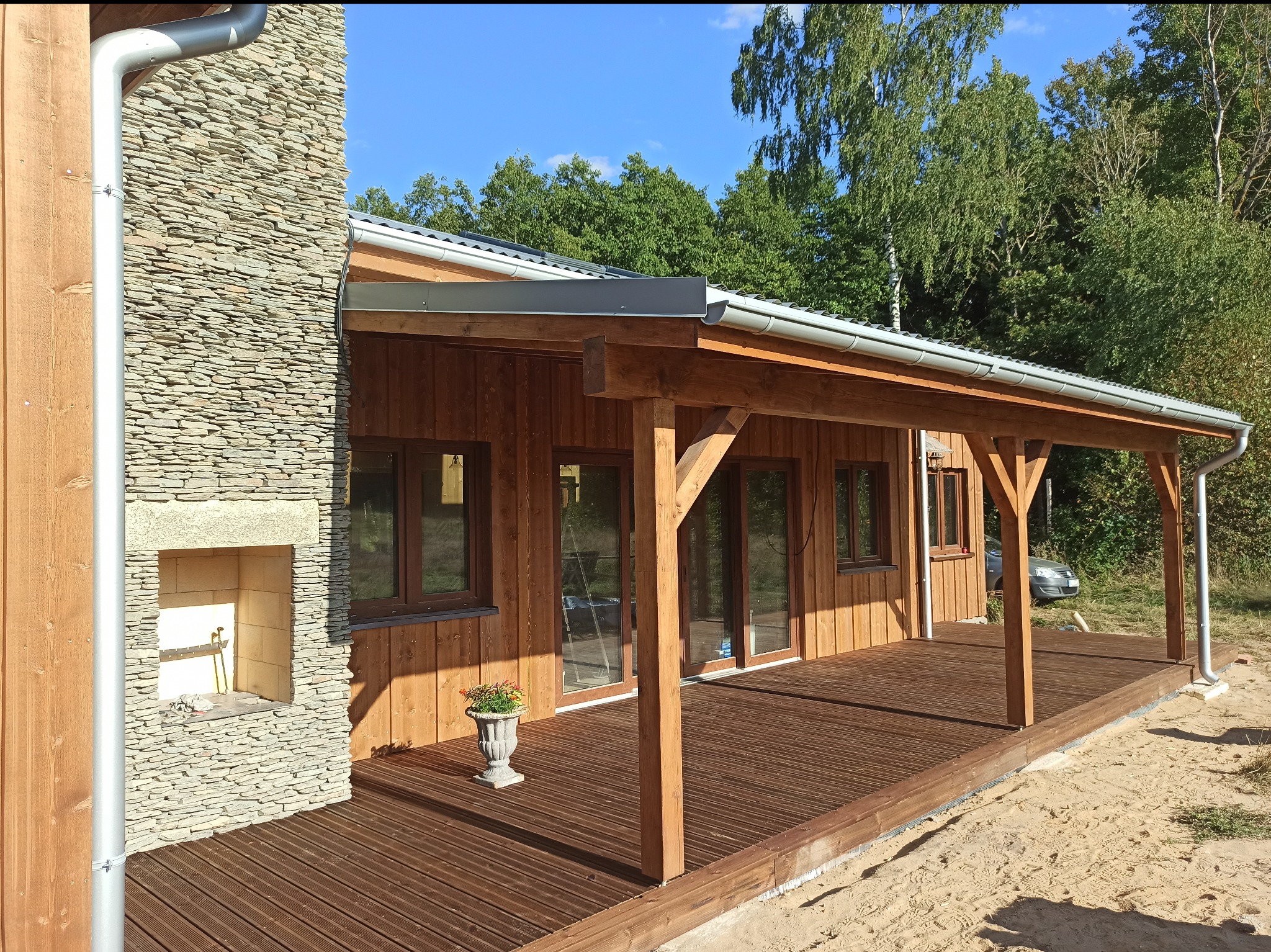 Drewniany dom z zadaszonym tarasem i kamiennym kominkiem. Na tarasie stoi ozdobna donica z kwiatami. W tle widoczne drzewa.