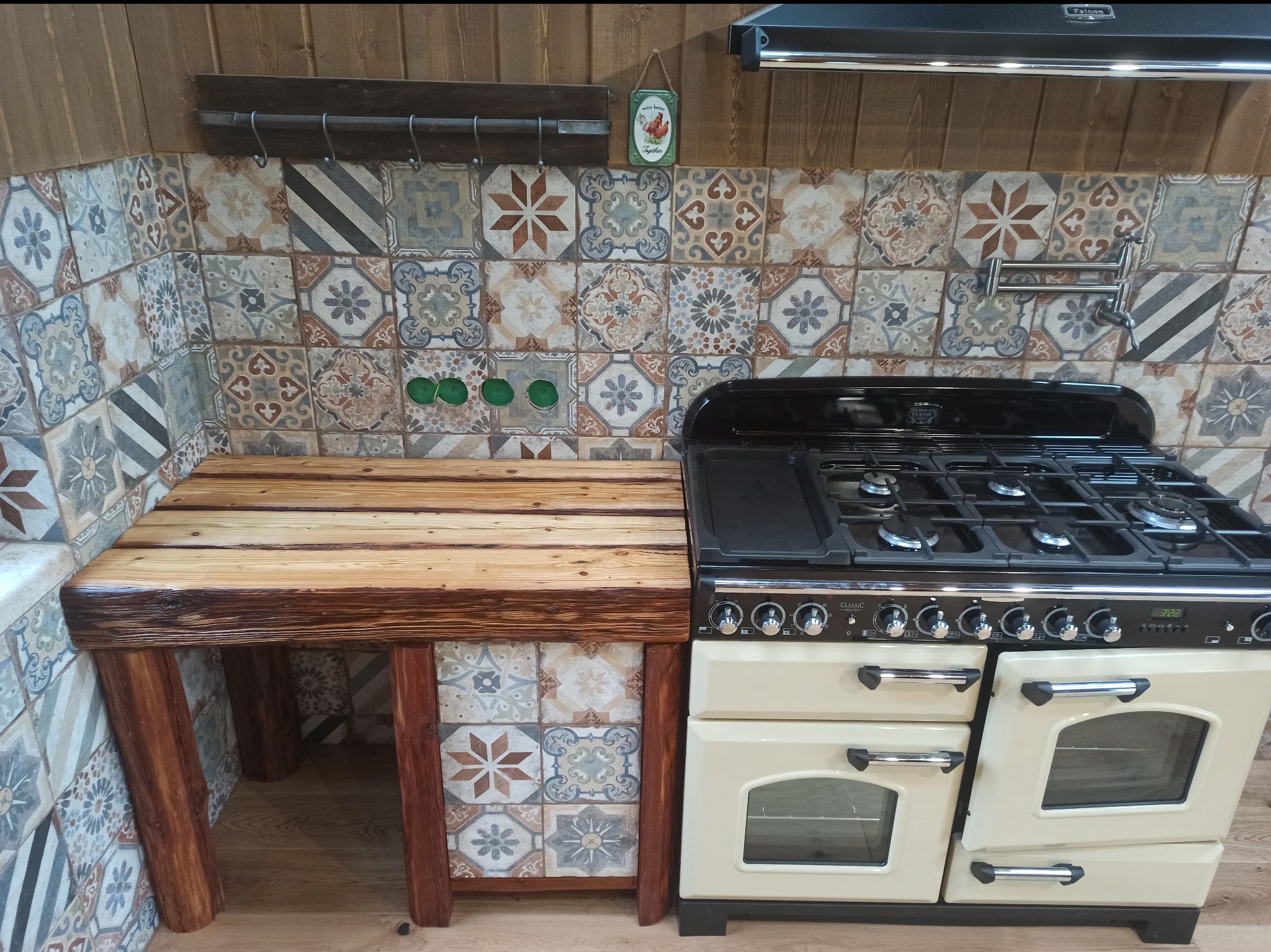 Rustykalna kuchnia: beżowa kuchenka gazowa Classic obok blatu z surowego drewna, tło z płytek w stylu patchwork. Wnętrze z drewnianymi panelami.