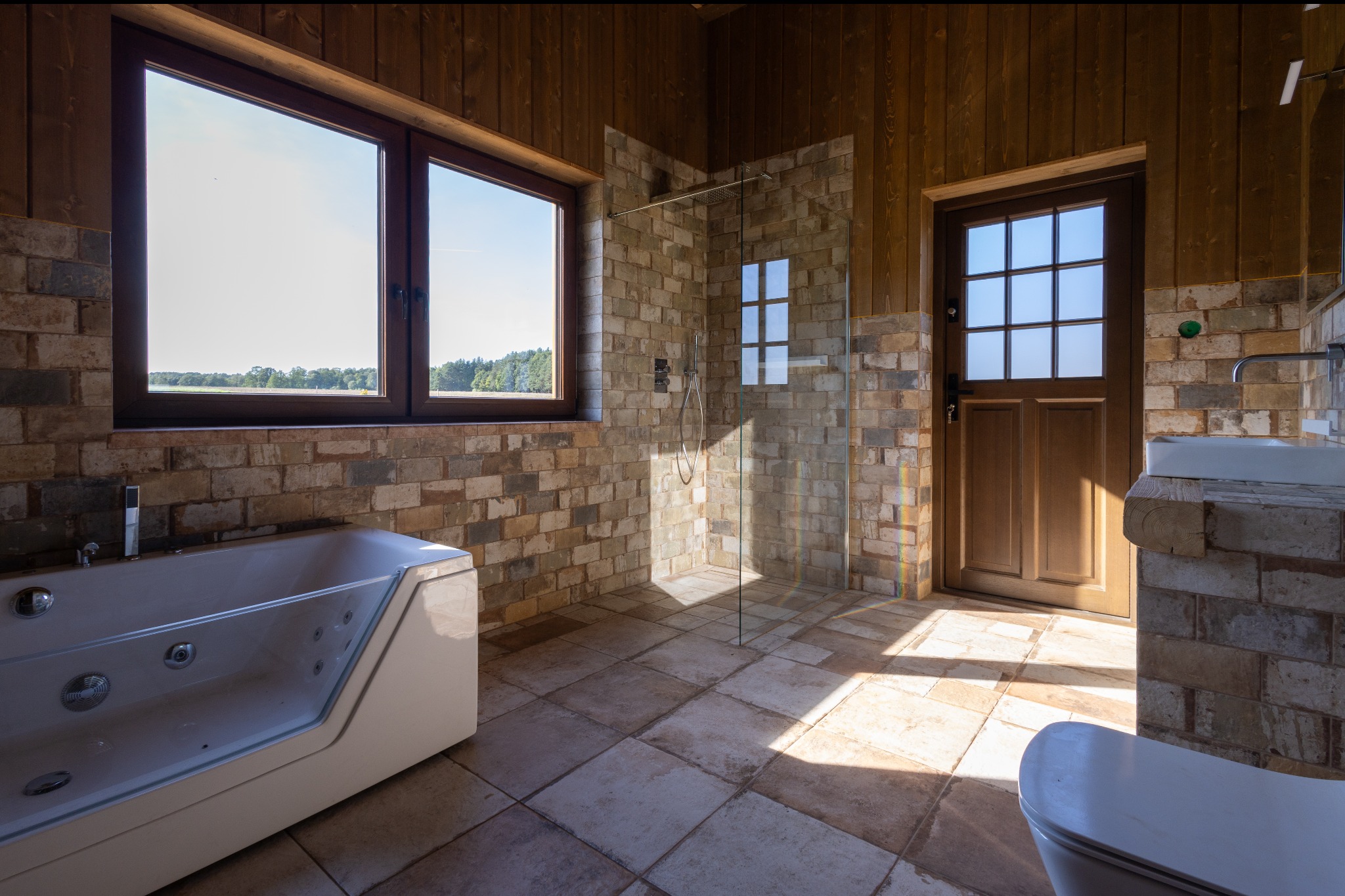 Rustykalna łazienka z kabiną prysznicową, wanną z hydromasażem i drewnianymi elementami wykończenia. Widok na jezioro przez okno.