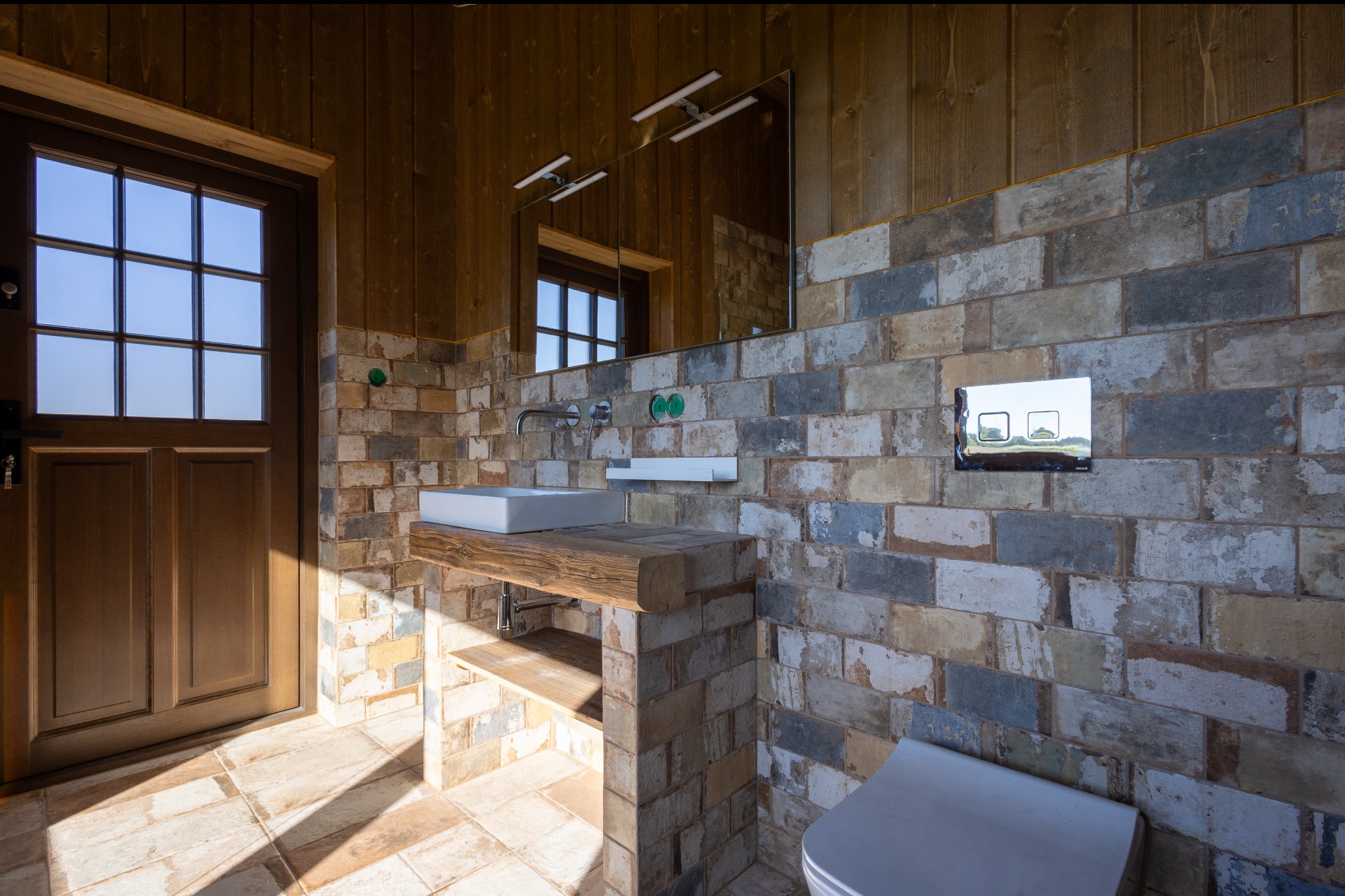 Rustykalna łazienka z drewnianym wykończeniem i ścianami z cegieł w odcieniach ziemi. Umywalka na drewnianym blacie w domu w Byczynie. Lustro i oświetlenie.