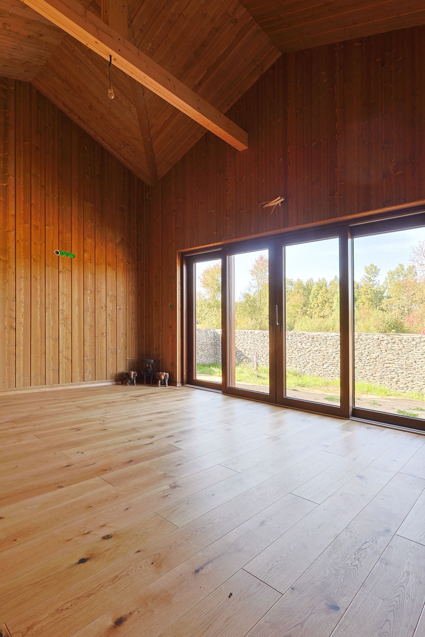 Wnętrze drewnianego domu z podłogą z jasnego drewna i dużymi oknami przesuwnymi z widokiem na kamienny murek i drzewa. Widoczne elementy instalacji elektrycznej.