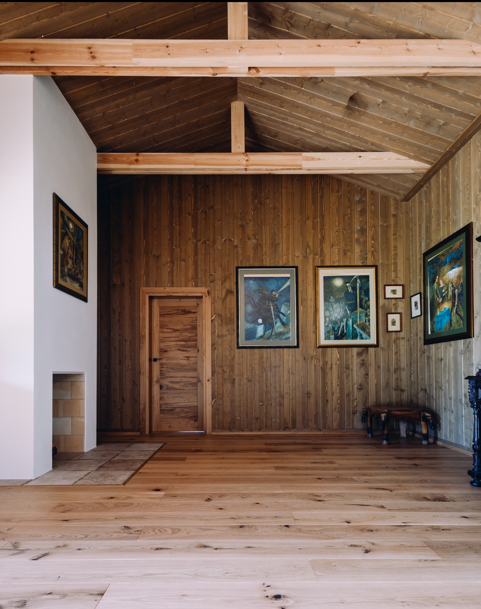 Drewniane wnętrze domu z boazerią, parkietem i belkami stropowymi. Na ścianach wiszą obrazy w ramach, a w rogu stoi ciemny, zdobiony mebel.