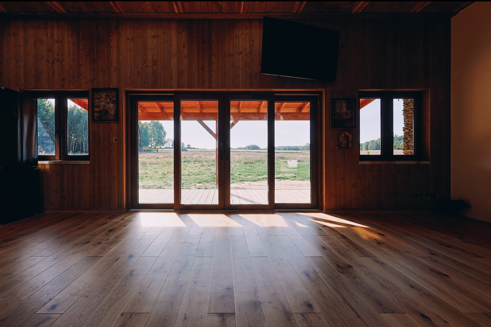 Wnętrze domu z drewnianą boazerią i podłogą. Widok na zewnątrz przez duże okna przesuwne. Na ścianie telewizor. Naturalne światło.