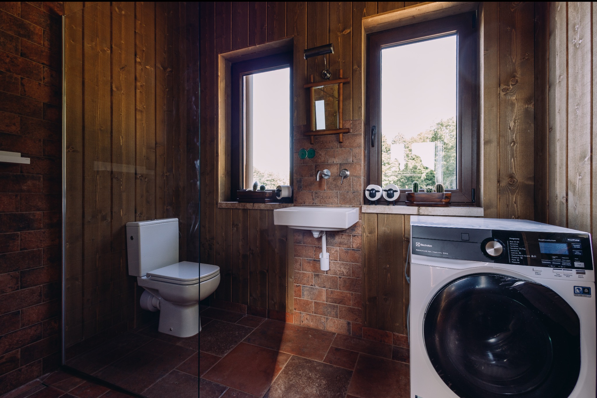 Rustykalna łazienka z drewnianymi ścianami i ceglaną zabudową umywalki. Widoczna toaleta, kabina prysznicowa i pralka Electrolux.