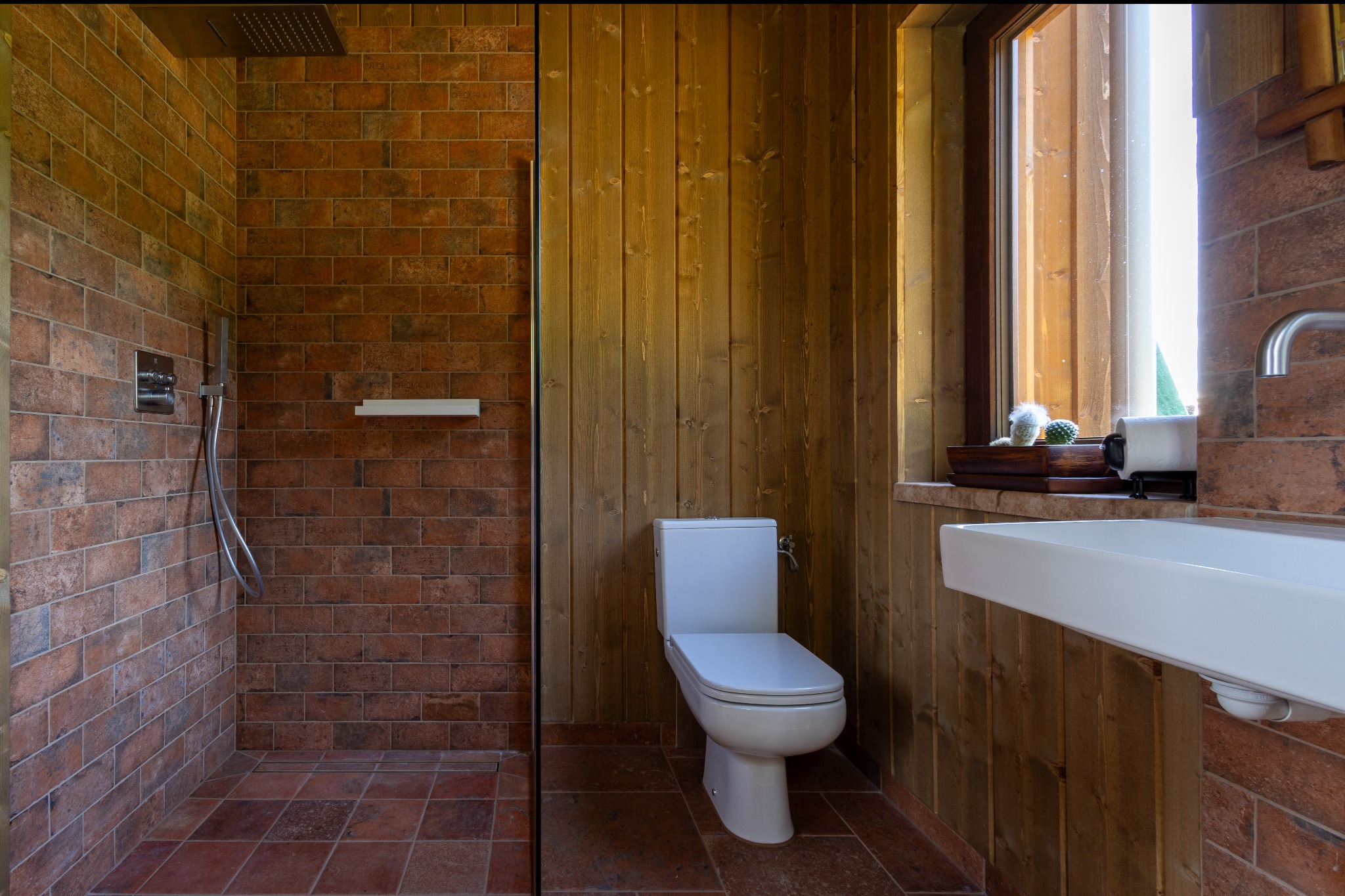 Rustykalna łazienka z kabiną prysznicową wyłożoną cegłą, drewnianą boazerią i oknem z dekoracjami. Umywalka i toaleta w minimalistycznym stylu.