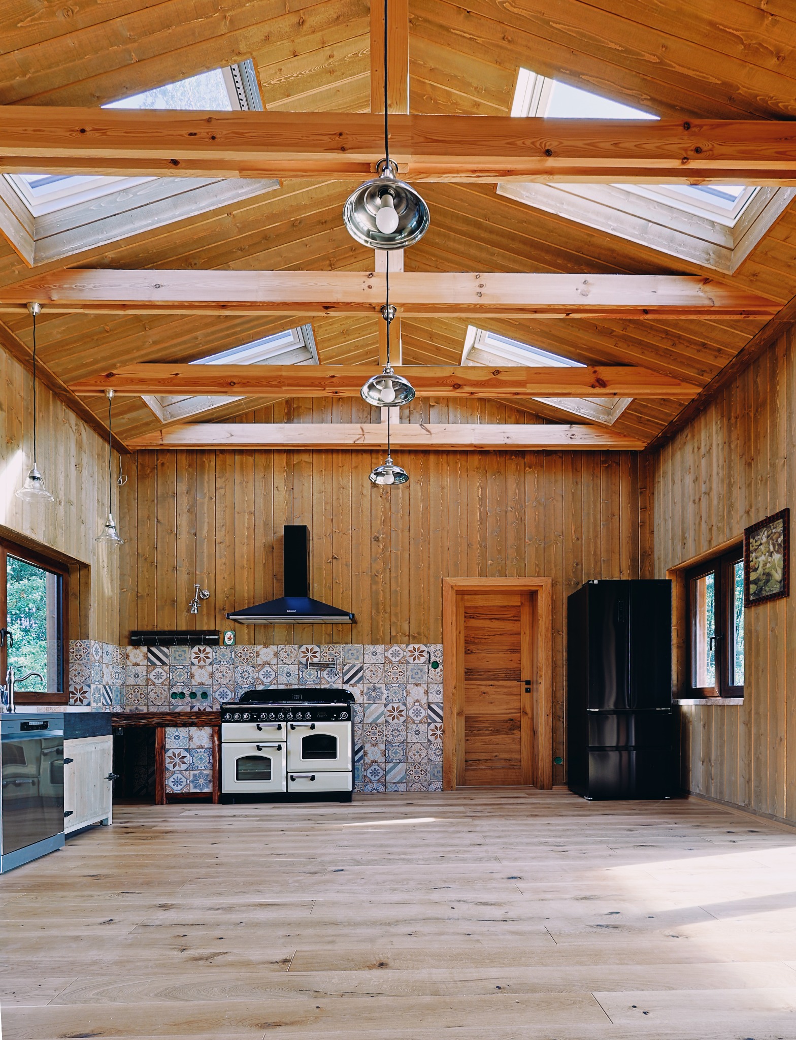 Wnętrze domu z drewnianą konstrukcją dachu i ścian, okna dachowe, kuchnia z kolorowymi płytkami, drewniana podłoga i wiszące lampy.