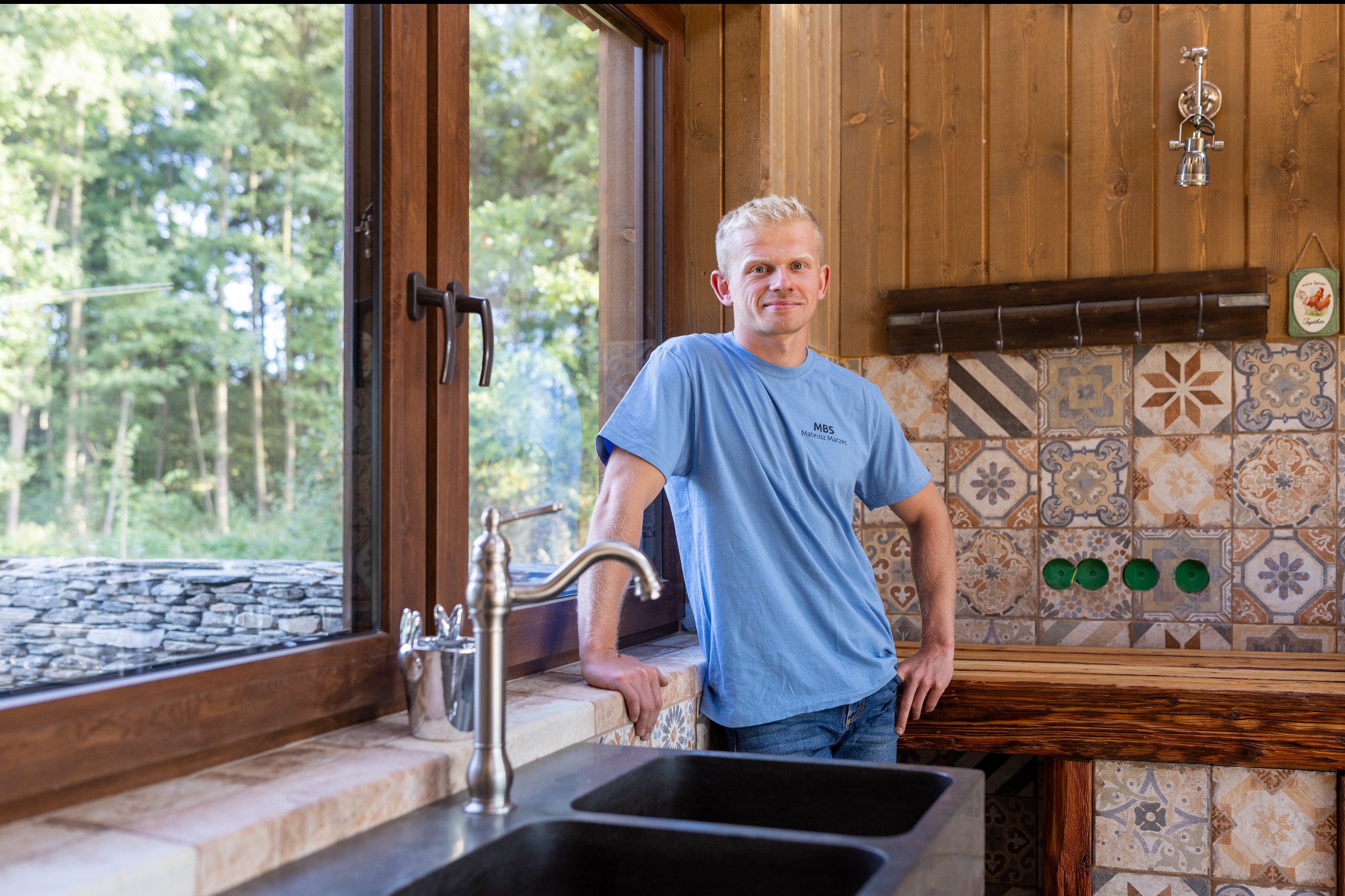 Wnętrze domu z drewnianymi ścianami i oknem z widokiem na zieleń. Mężczyzna w niebieskiej koszulce opiera się o blat przy zlewie, z tyłu ściana z dekoracyjnymi płytkami.