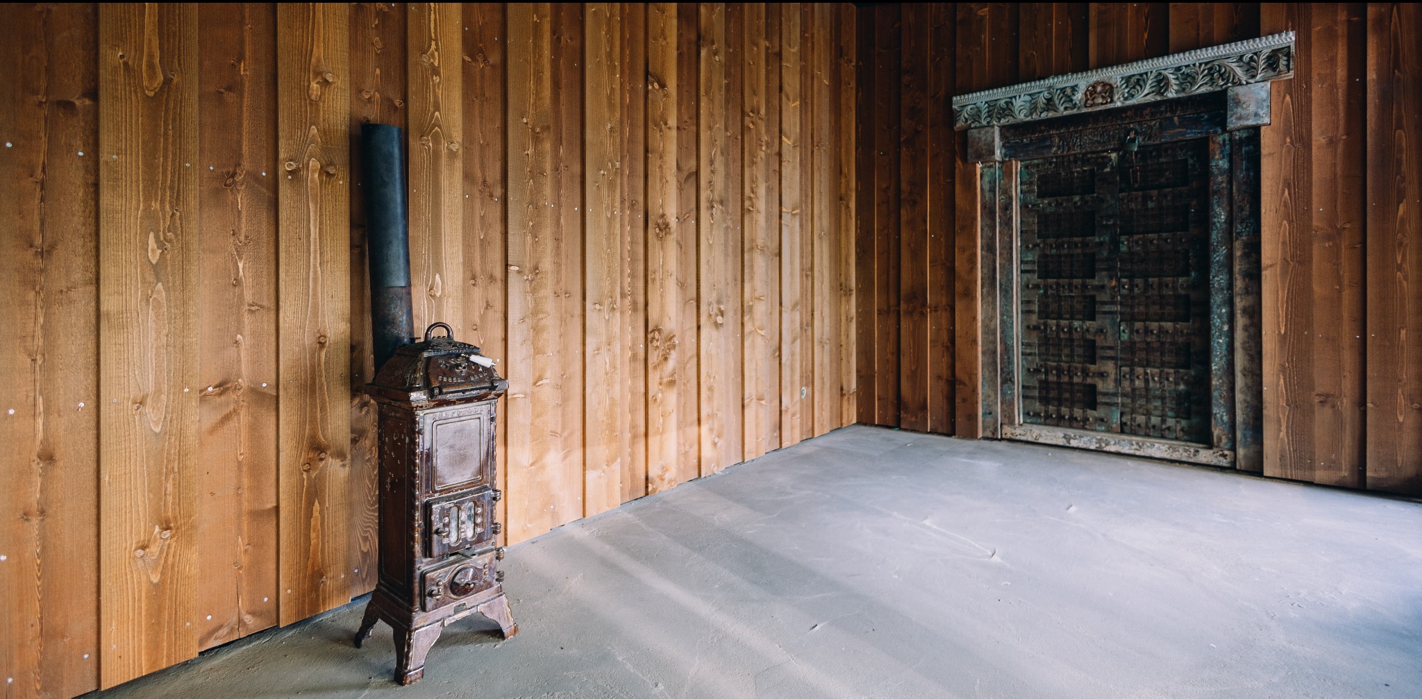 Wnętrze z boazerią i piecem typu koza. Ciemne drewno na ścianach i podłodze. W tle ozdobne, ciemne drzwi. Całość w klimacie retro.