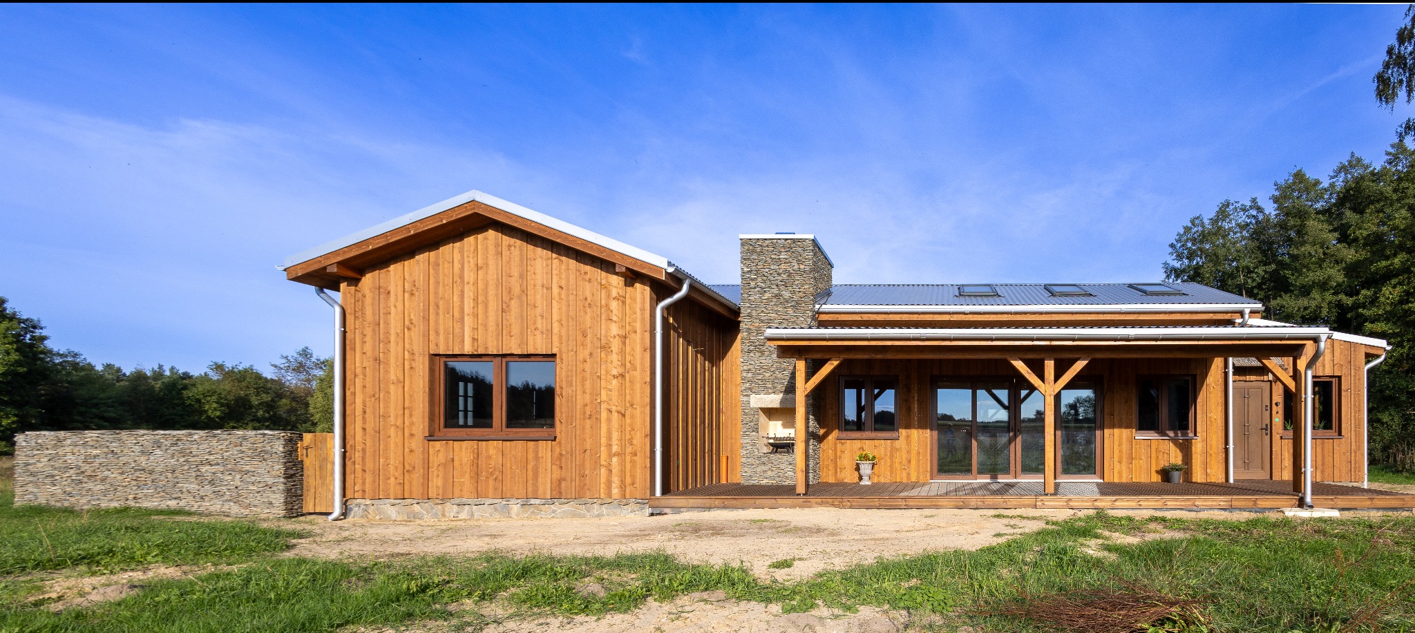 Parterowy dom z drewna o jasnej elewacji, z kamiennym kominem i zadaszonym tarasem. Widoczne okna, drzwi i fragment kamiennego ogrodzenia.