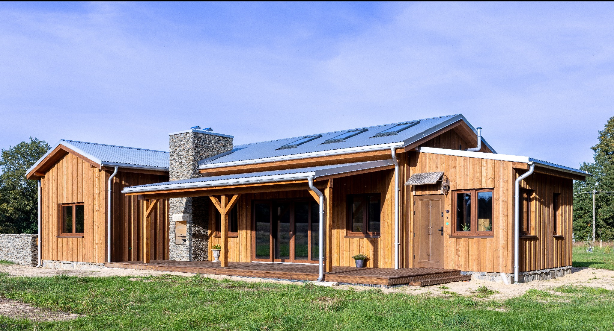 Drewniany dom z metalowym dachem i kamiennym kominem w stylu rustykalnym. Widoczny zadaszony taras i starannie wykończone detale.