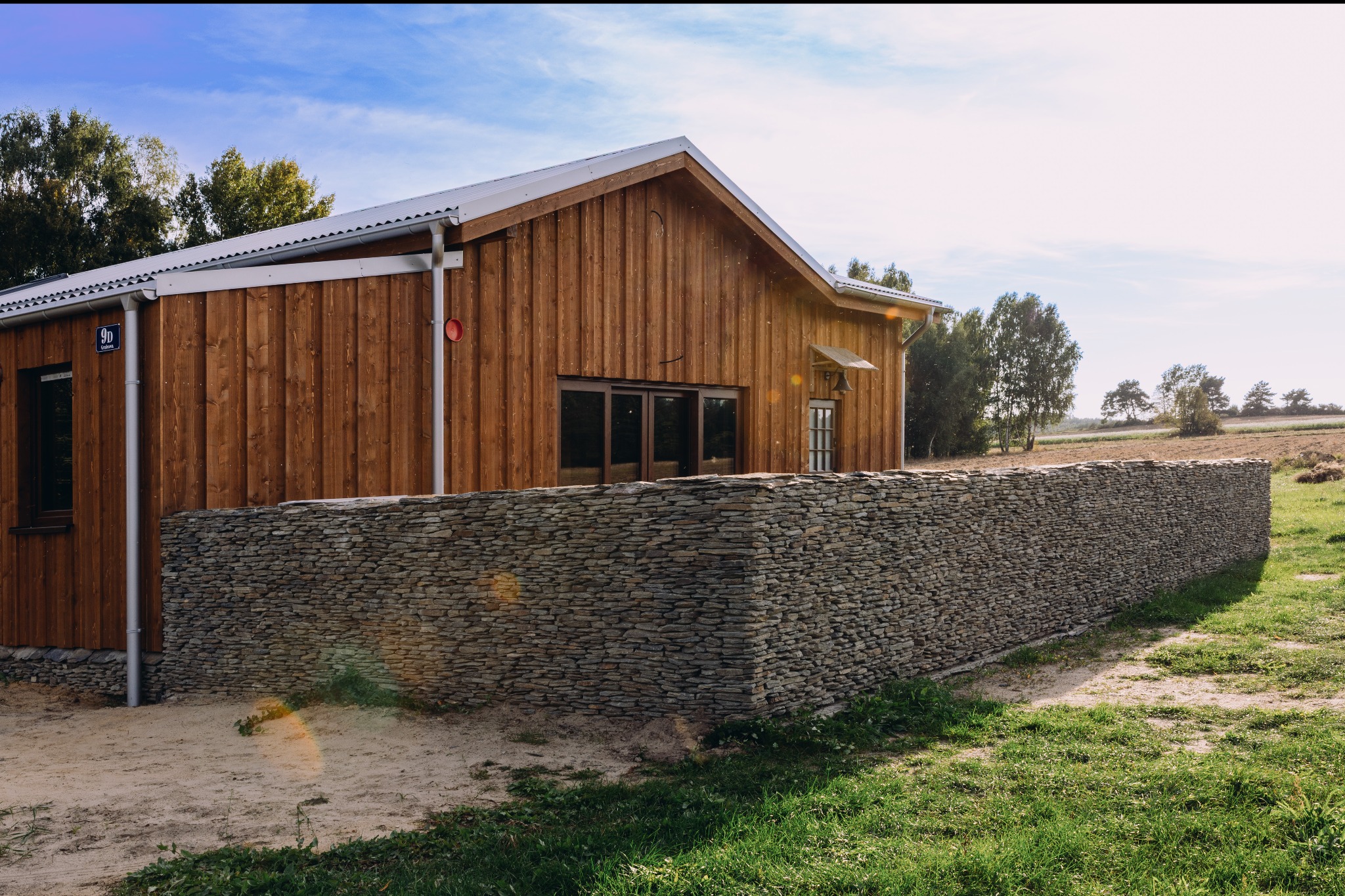 Drewniany dom z charakterystyczną fasadą i kamiennym ogrodzeniem. Widoczny numer domu '9D'. Ujęcie w słoneczny dzień, z fragmentem zieleni i drzew w tle.