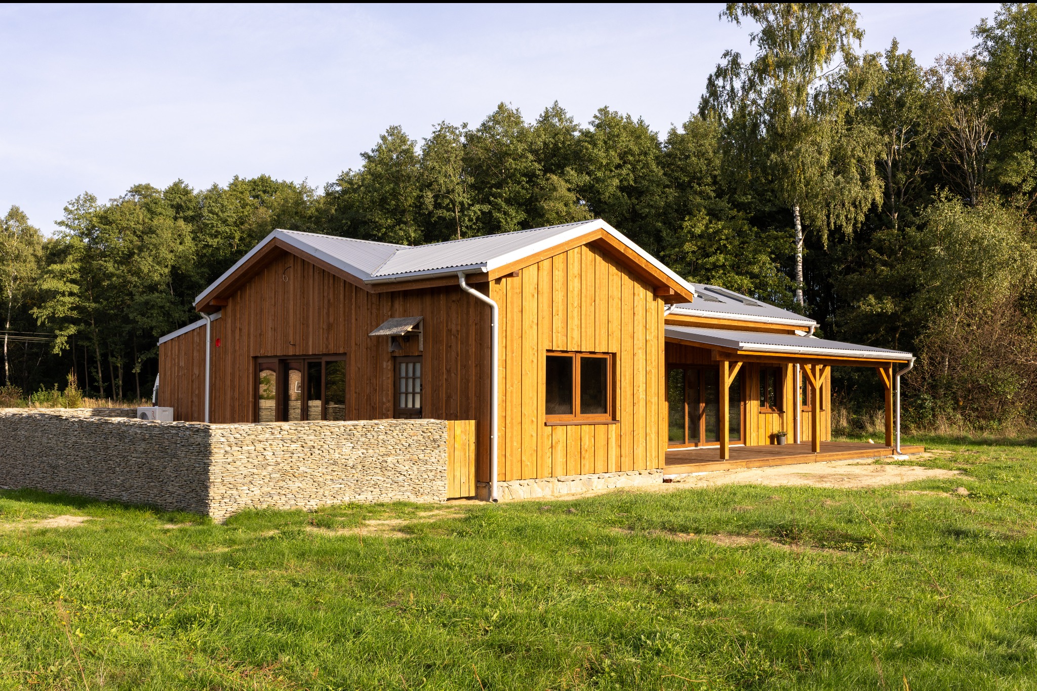 Drewniany dom z szarym dachem i kamiennym ogrodzeniem na tle zieleni. Widoczne okna, drzwi i zadaszony taras. Jasne drewno elewacji, naturalne światło.