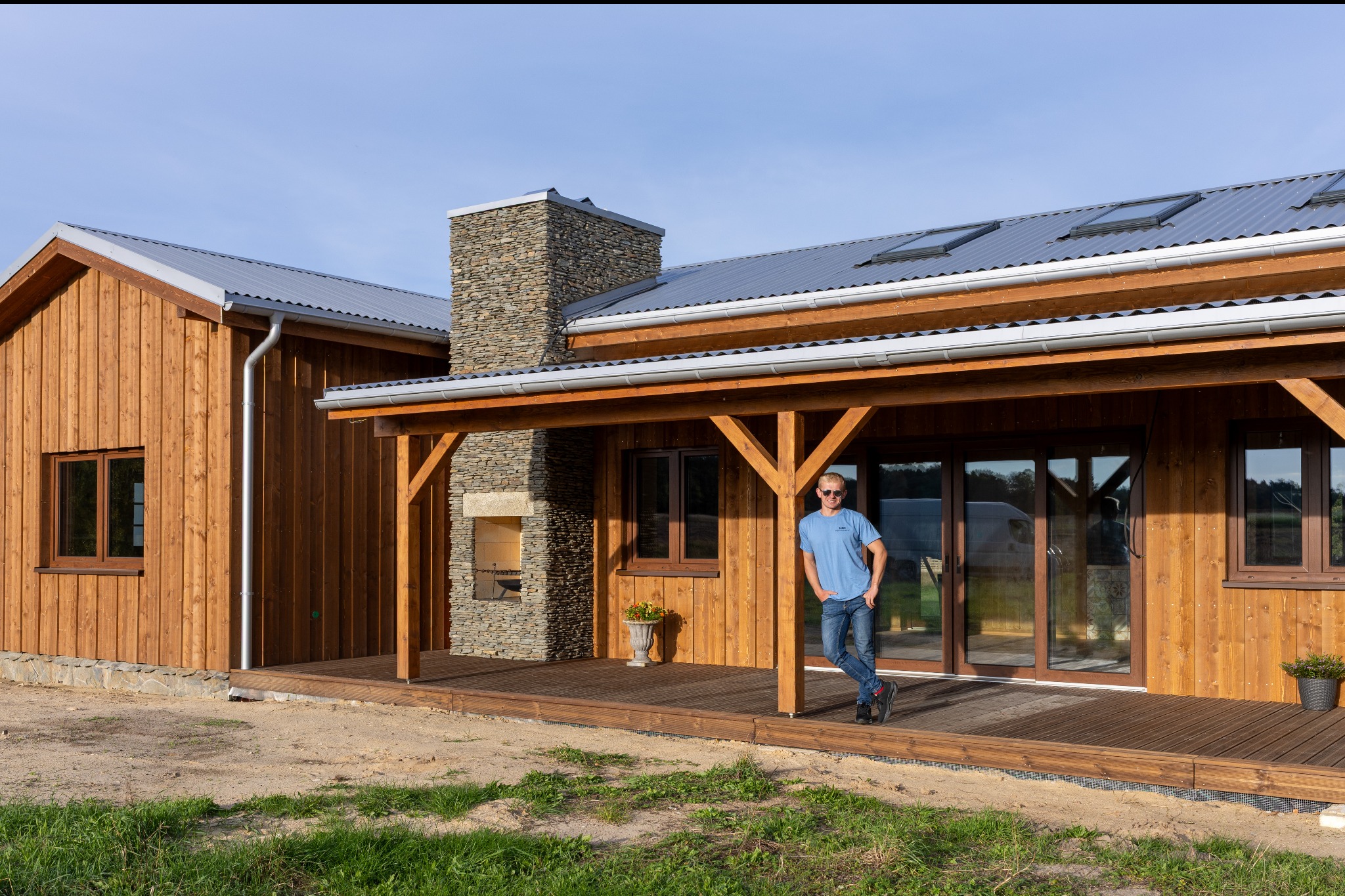 Drewniany dom z kamiennym kominem i zadaszonym tarasem, na którym stoi mężczyzna. Dach pokryty blachą, okna z brązowymi ramami. Całość w naturalnym otoczeniu.