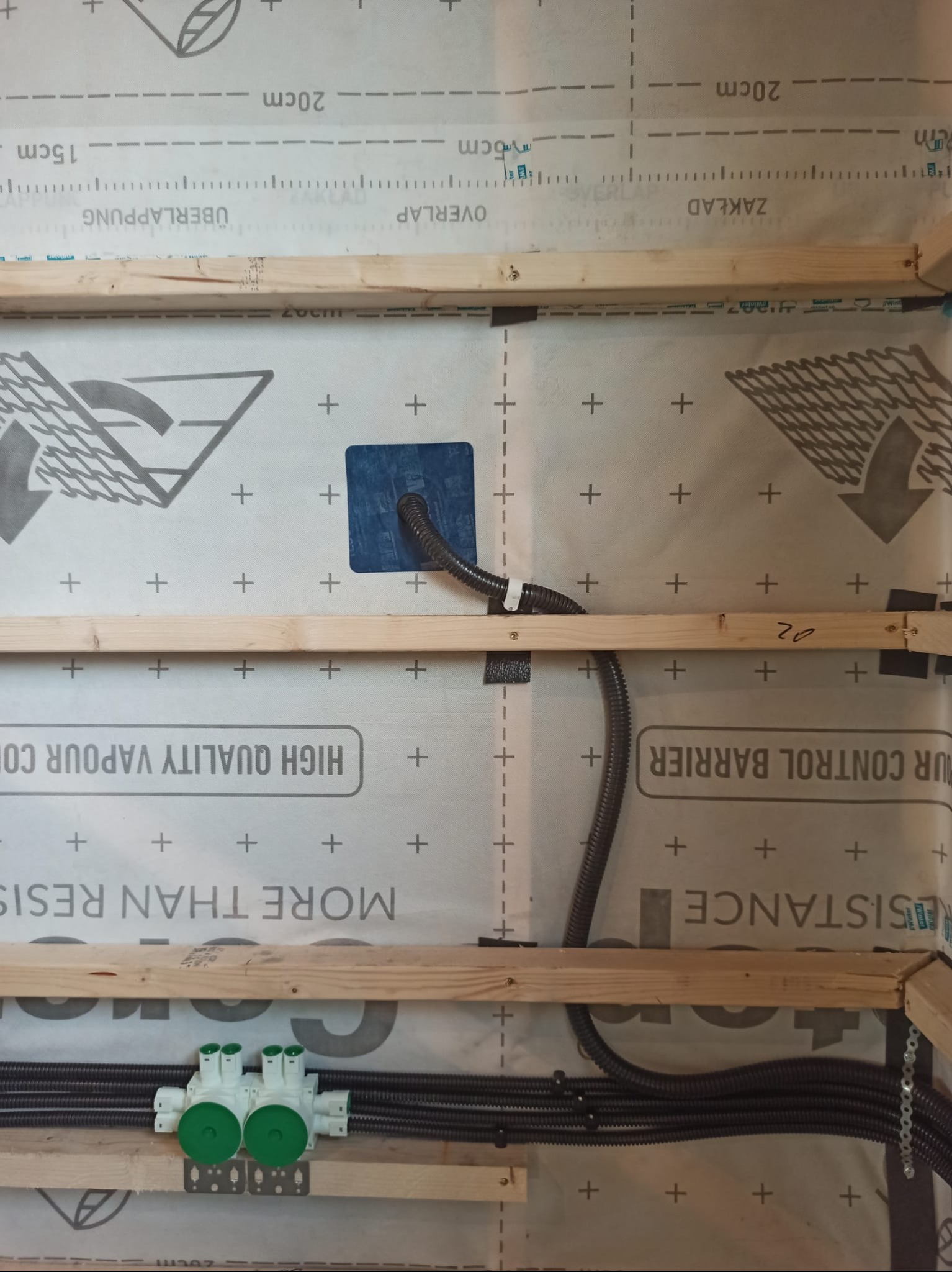 Fragment ściany szkieletowej z widoczną instalacją elektryczną i wodną. Elementy drewniane konstrukcji, folia paroizolacyjna z nadrukiem. Detal instalacji wewnątrz ściany.