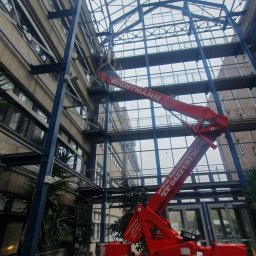 Masz Problem Zadzwoń Usuniemy Go 5*1*9 3*5*3 3*2*8 - Czerwony podnośnik koszowy marki LEO na gąsienicach, z operatorem w koszu, używany do prac na wysokości wewnątrz budynku z przeszklonym dachem i metalową konstrukcją szkieletową.