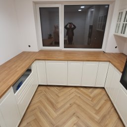 Masz Problem Zadzwoń Usuniemy Go 5*1*9 3*5*3 3*2*8 - Jasna kuchnia z białymi szafkami i drewnianym blatem, parkiet ułożony w jodełkę, okno z odbiciem osoby robiącej zdjęcie
