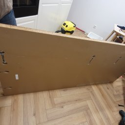 Masz Problem Zadzwoń Usuniemy Go 5*1*9 3*5*3 3*2*8 - Montaż płyty meblowej z otworami na łączniki mimośrodowe w trakcie prac wykończeniowych, widoczny odkurzacz i fragmenty innych mebli.