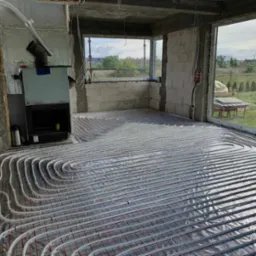 Surowy pokój z widocznym systemem ogrzewania podłogowego rozłożonym na folii izolacyjnej, kominek w stanie surowym, widok przez okno na zewnątrz.