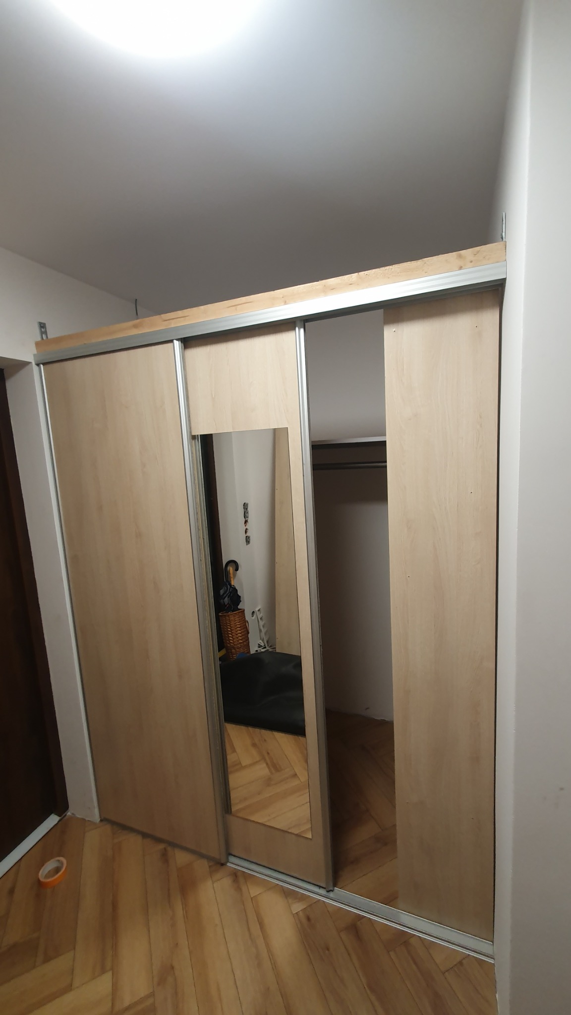 Zamontowana szafa wnękowa z przesuwnymi drzwiami z jasnego drewna i lustrem, widoczna podłoga z paneli ułożonych w jodełkę.