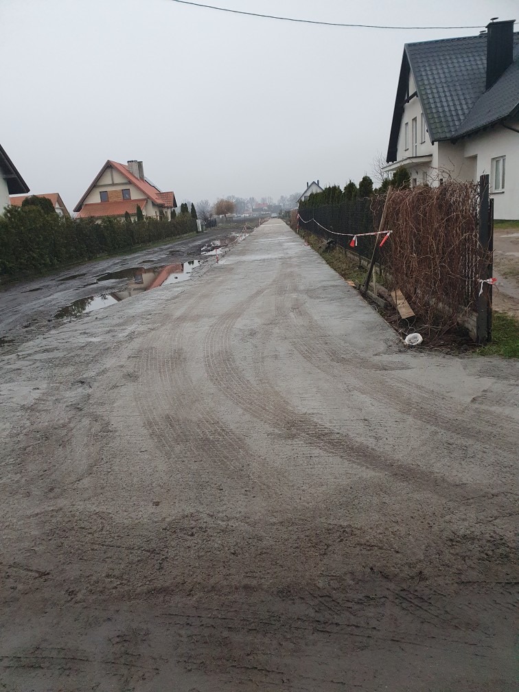 Świeżo wylany, mokry beton na podjeździe w zabudowie jednorodzinnej, widoczne ślady opon, zabezpieczony taśmami ostrzegawczymi, pochmurny dzień.