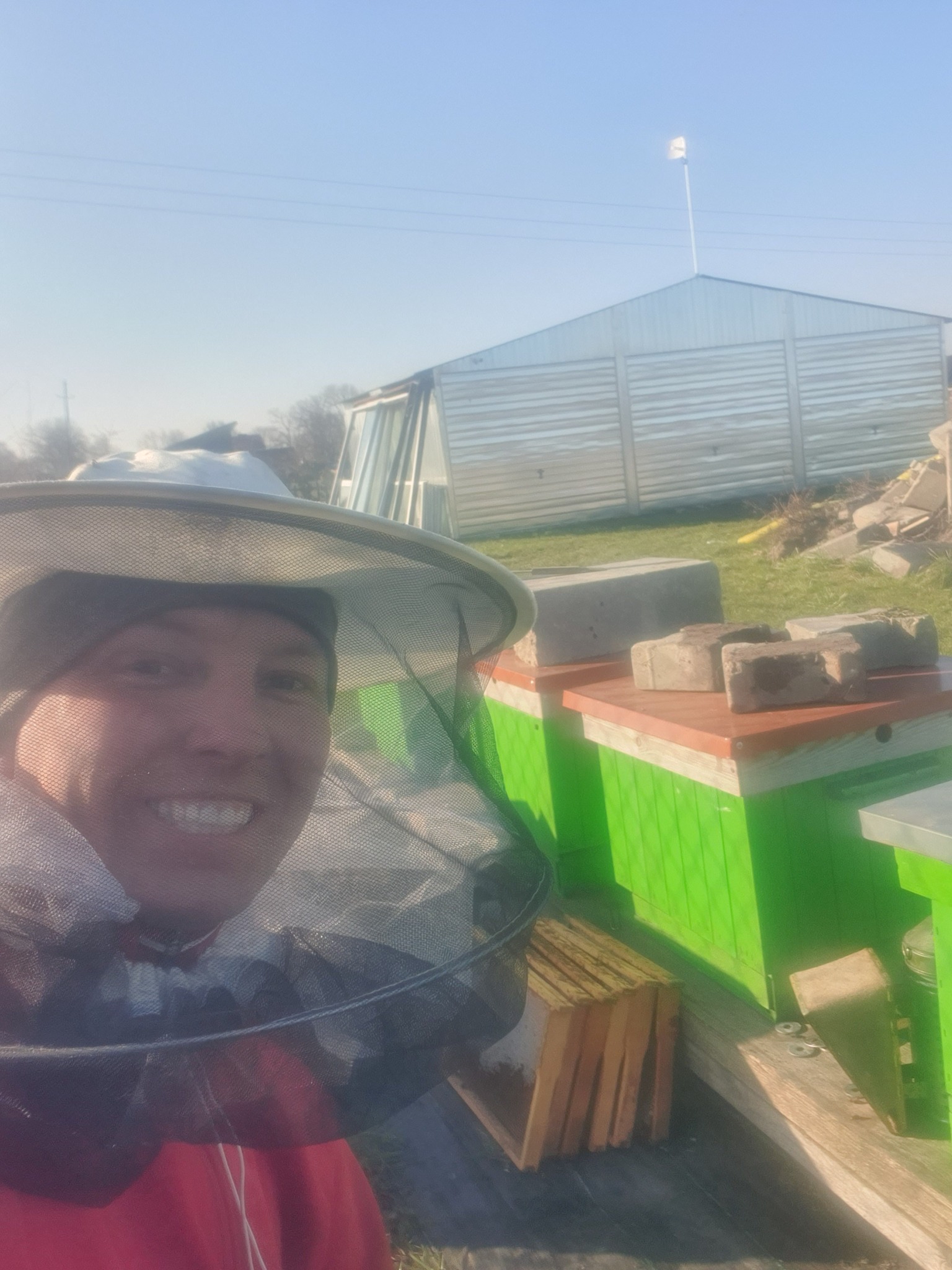 Uśmiechnięty pszczelarz w siatce ochronnej na tle uli pomalowanych na zielono, z cegłami na wierzchu, oraz blaszanego garażu w słoneczny dzień.