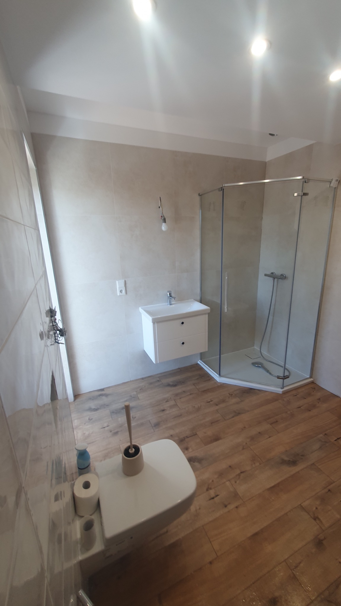 Jasna łazienka po remoncie z drewnopodobną podłogą, kabiną prysznicową i podwieszaną szafką z umywalką. Na ścianie widoczny włącznik światła i wisząca żarówka bez klosza.