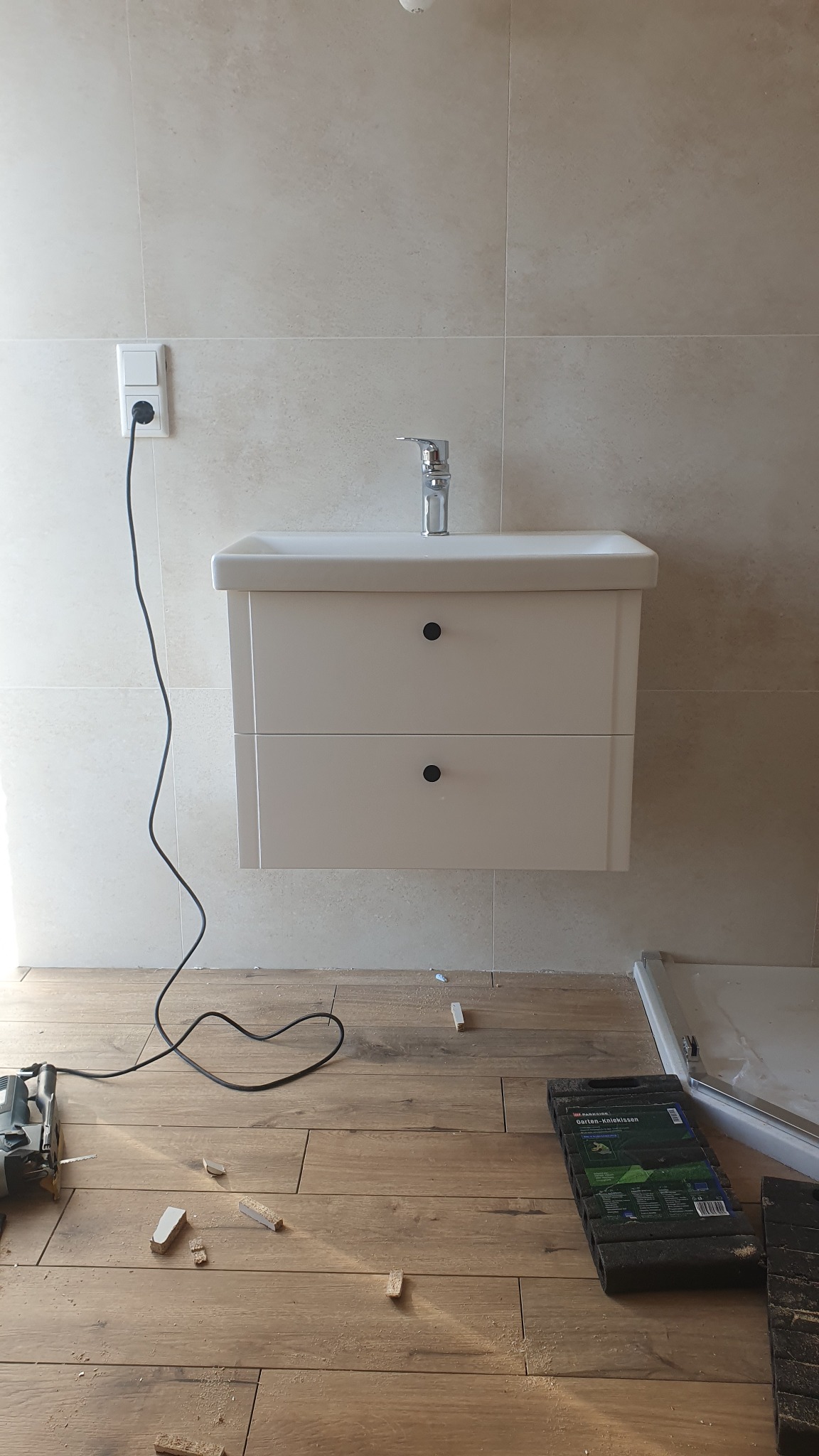 Montaż umywalki z szafką w łazience w trakcie prac wykończeniowych, widoczne narzędzia i fragmenty podłogi, jasne płytki na ścianie.