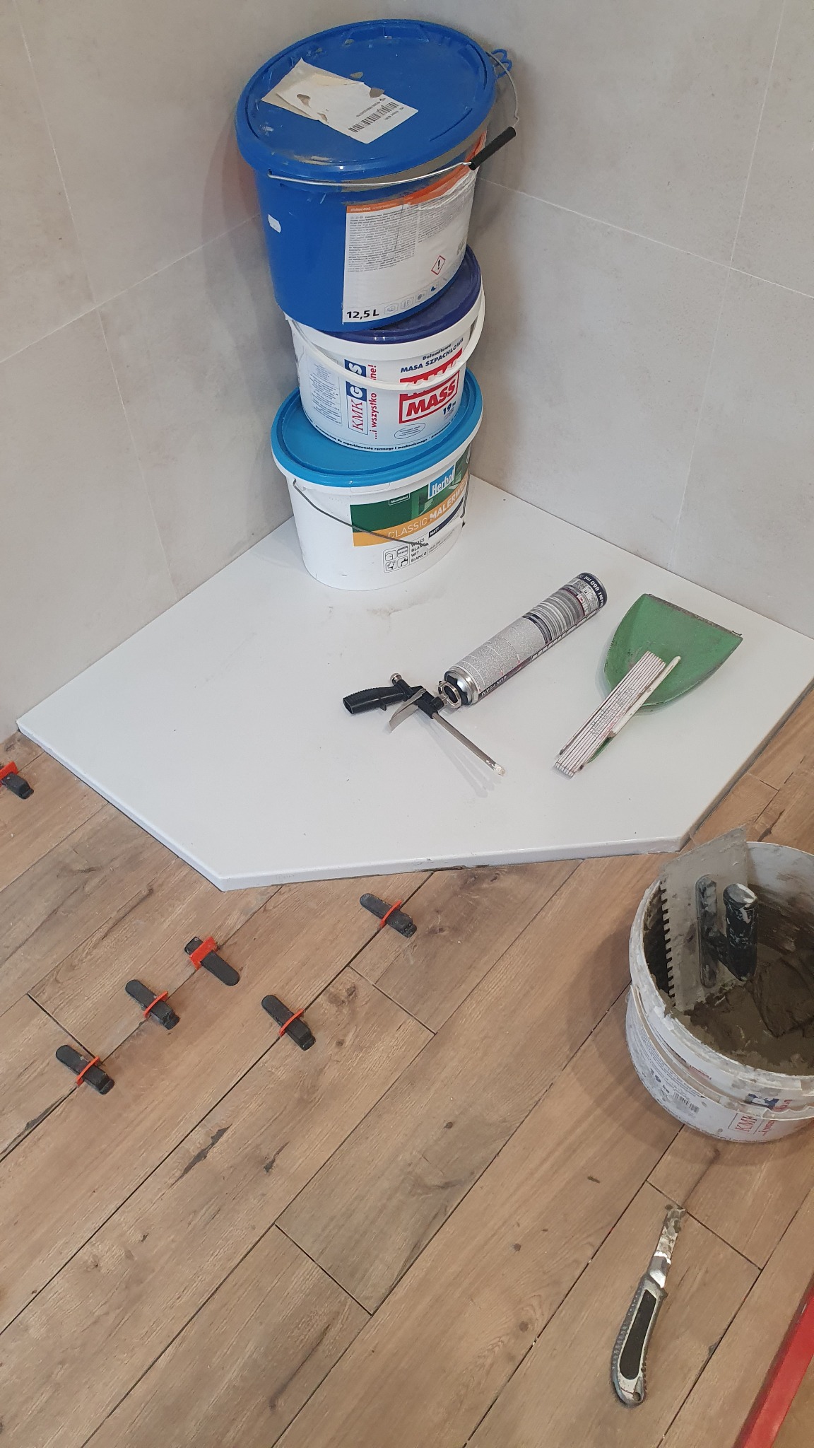 Układanie drewnopodobnych płytek w rogu łazienki, widoczne narzędzia glazurnicze, wiadro z zaprawą, pistolet do silikonu, poziomica, kliny i krzyżyki dystansowe, trzy wiadra farb i mas szpachlowych...