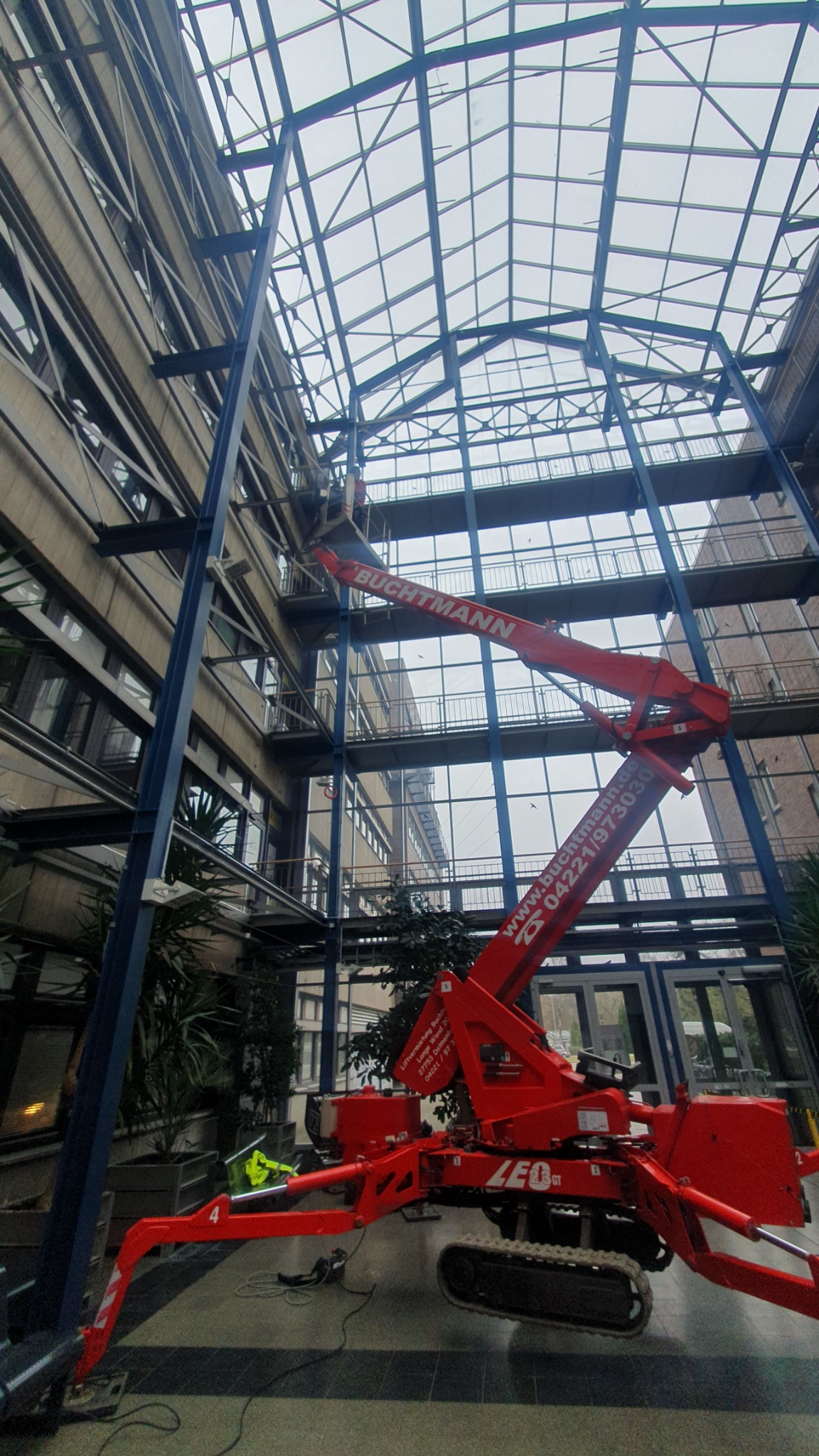 Czerwony podnośnik koszowy marki LEO na gąsienicach, z operatorem w koszu, używany do prac na wysokości wewnątrz budynku z przeszklonym dachem i metalową konstrukcją szkieletową.