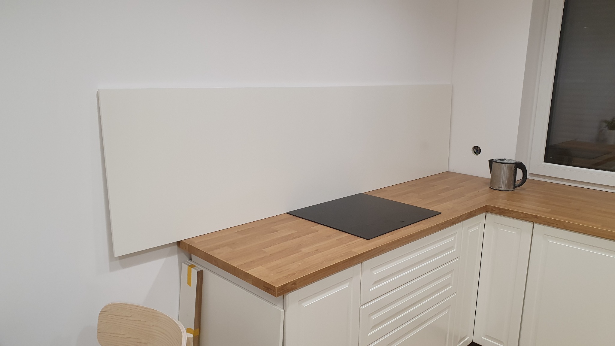 Biała kuchnia z drewnianym blatem, czarną płytą indukcyjną i srebrnym czajnikiem elektrycznym przy oknie. Na ścianie biała płyta ochronna. Widoczny otwór w ścianie po instalacji elektrycznej.