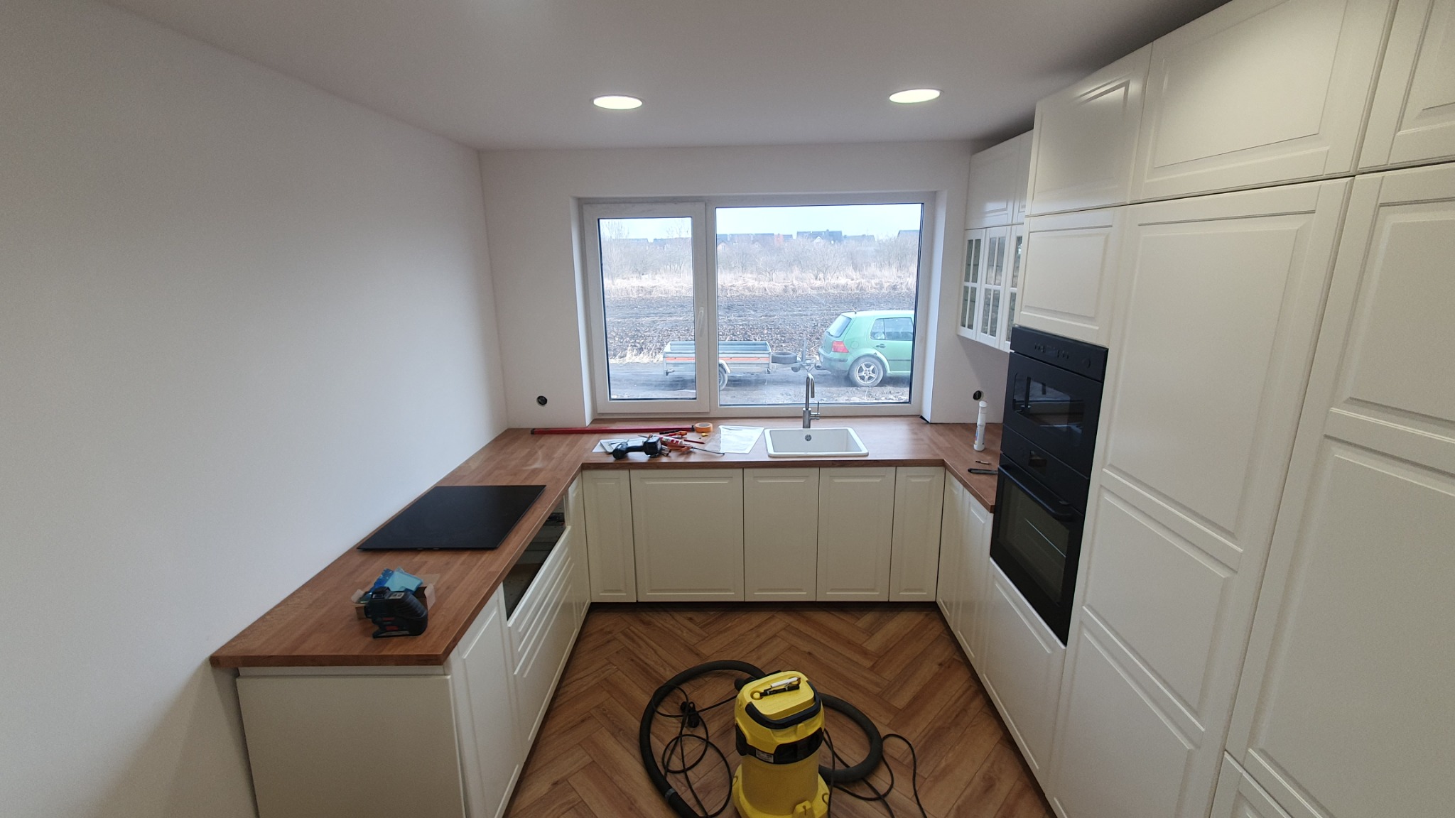 Nowa kuchnia z białymi szafkami i drewnianym blatem, widoczny zlewozmywak, płyta indukcyjna, piekarnik w zabudowie i odkurzacz na parkiecie w jodełkę.