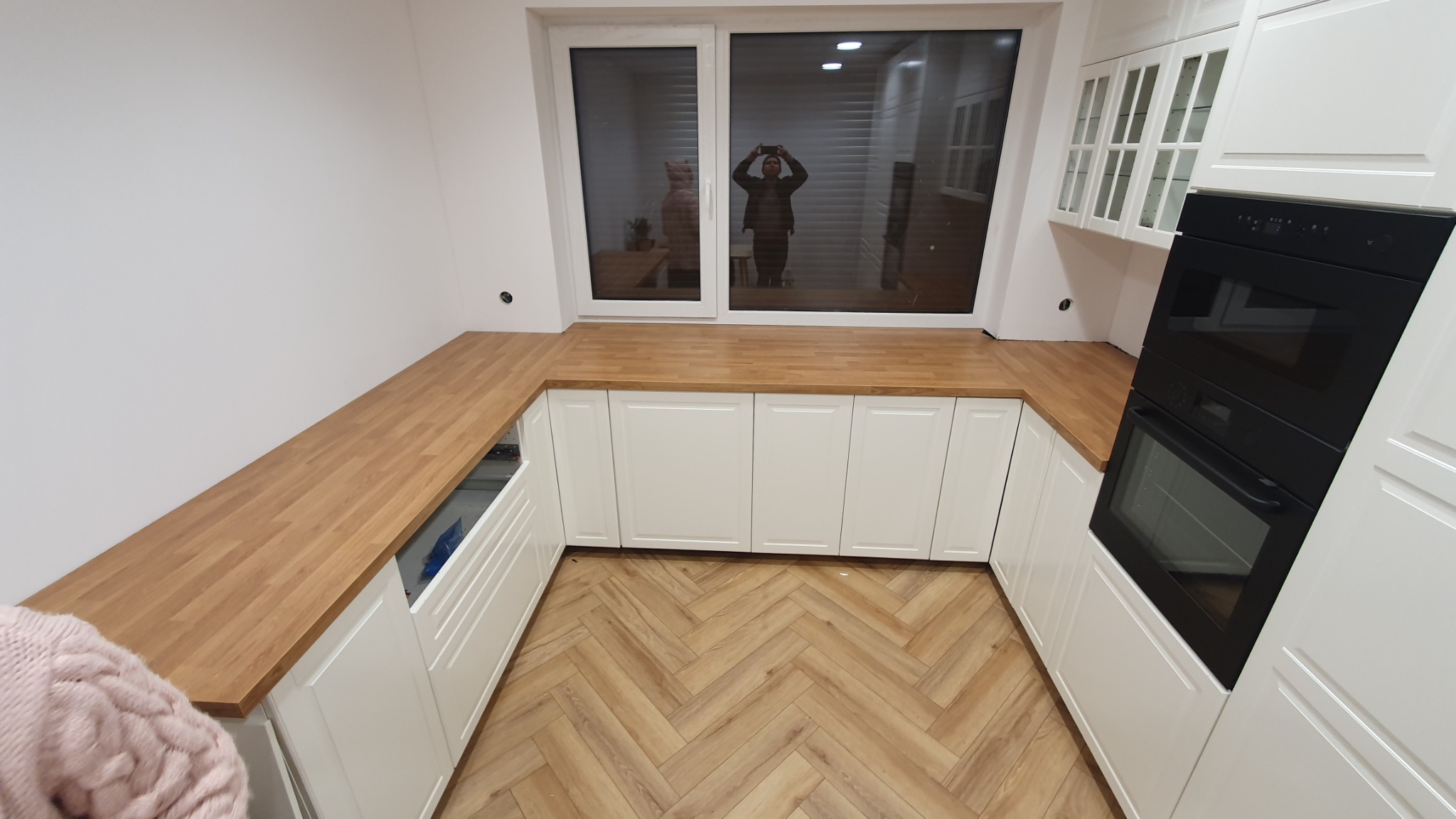 Jasna kuchnia z białymi szafkami i drewnianym blatem, parkiet ułożony w jodełkę, okno z odbiciem osoby robiącej zdjęcie