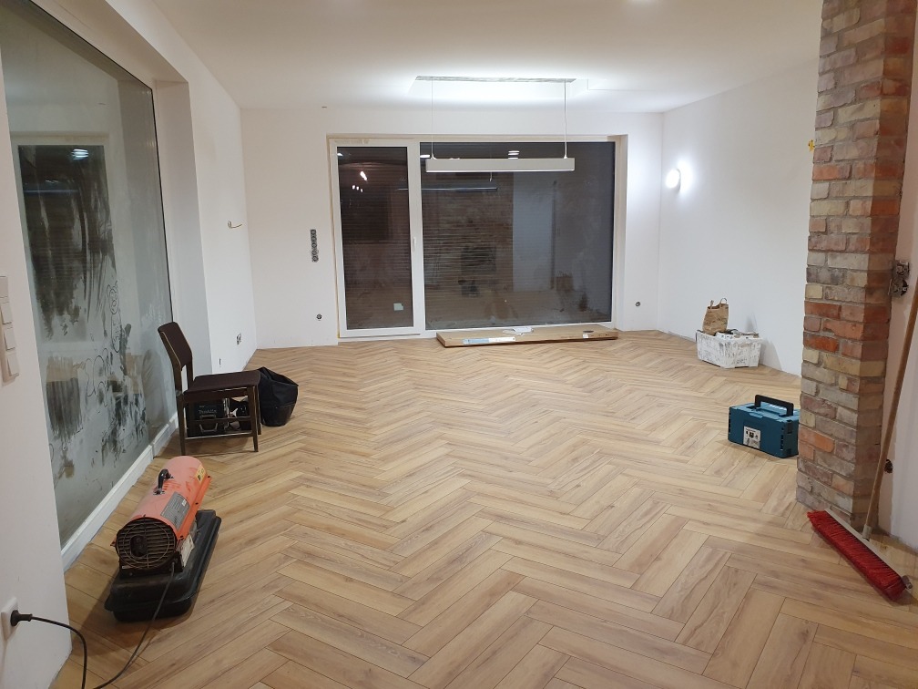 Nowo ułożony parkiet w jodełkę w przestronnym, jasnym pokoju z dużymi oknami, widoczny grzejnik budowlany, krzesło i narzędzia.