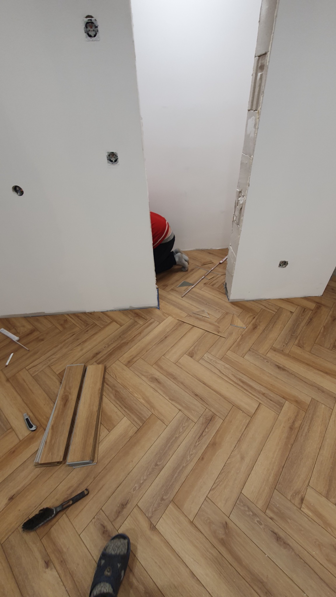 Układanie parkietu w jodełkę w trakcie prac wykończeniowych, widoczny pracownik przycinający panele w przejściu, obok leżą panele, miotła i nóż do cięcia