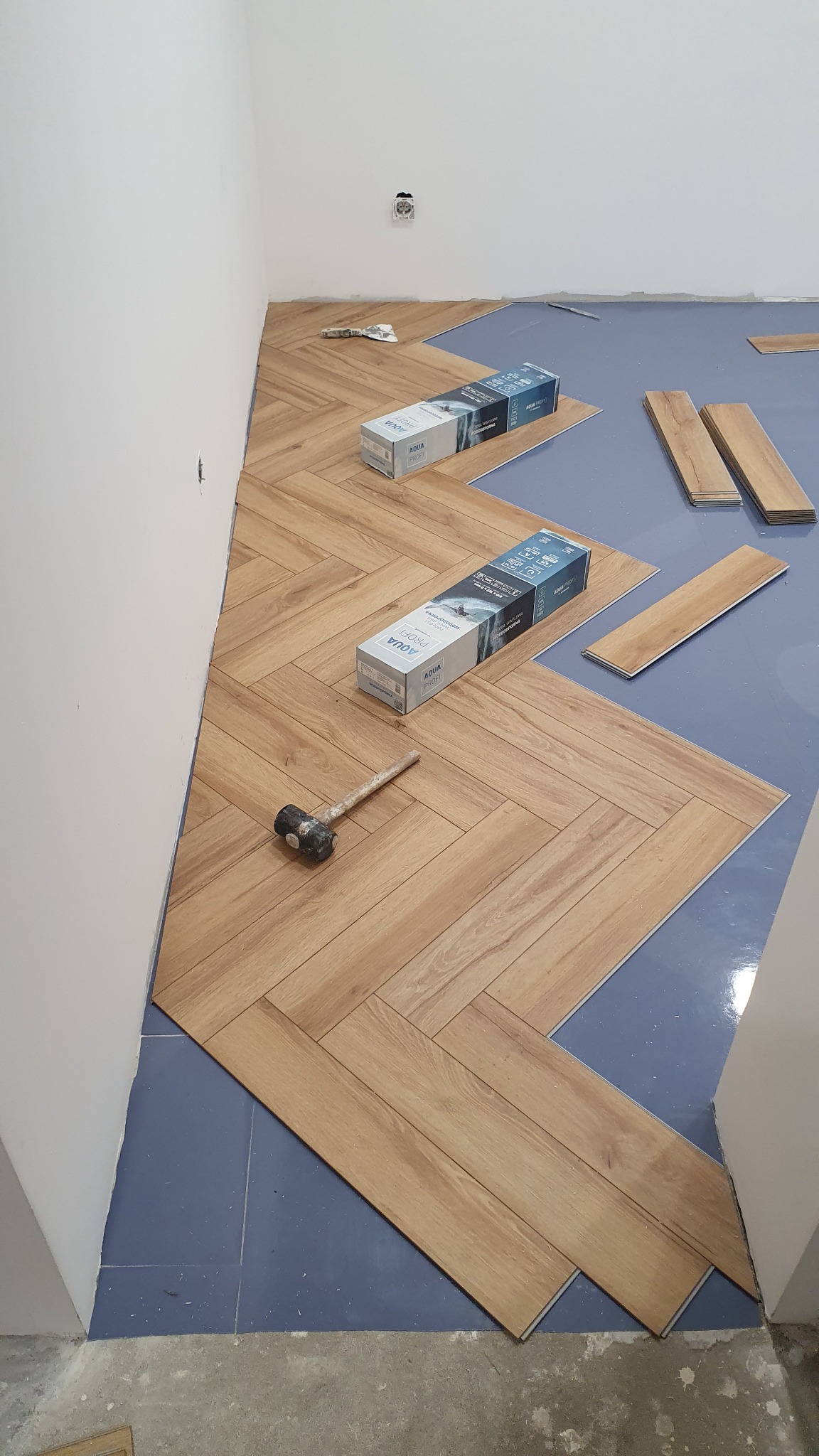 Układanie parkietu w jodełkę, widoczne paczki paneli Aqua Profi, gumowy młotek i fragment podłoża zabezpieczonego folią.