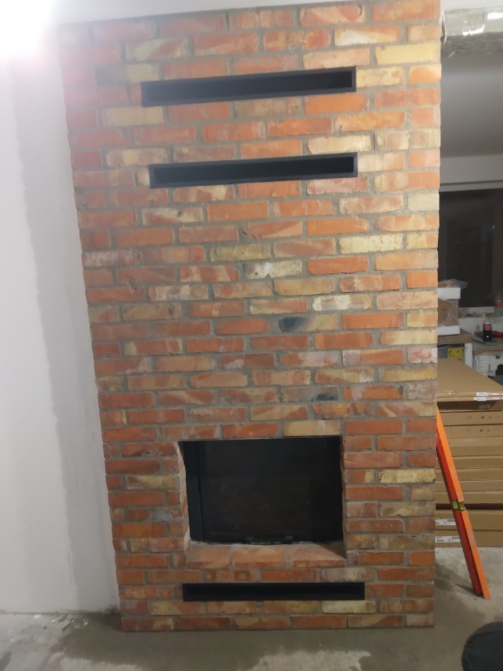 Murowany kominek z czerwonej cegły z poziomymi szczelinami wentylacyjnymi i czarnym wkładem kominkowym, widoczny poziomica oparta o ścianę, surowy beton na podłodze.