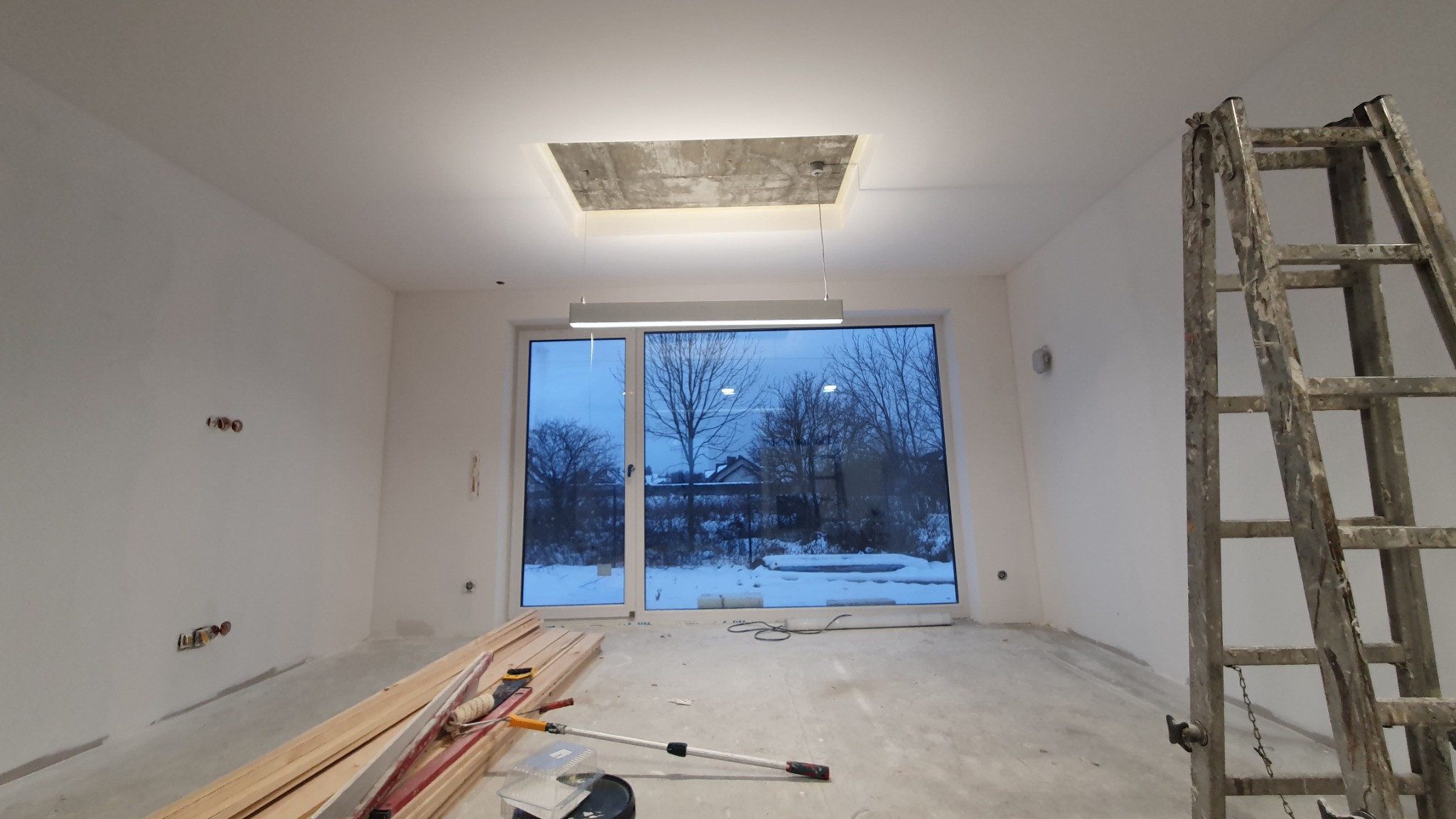 Wnętrze pokoju w trakcie prac wykończeniowych; widoczne przygotowane deski podłogowe, drabina malarska, oraz surowy betonowy element oświetleniowy w suficie.