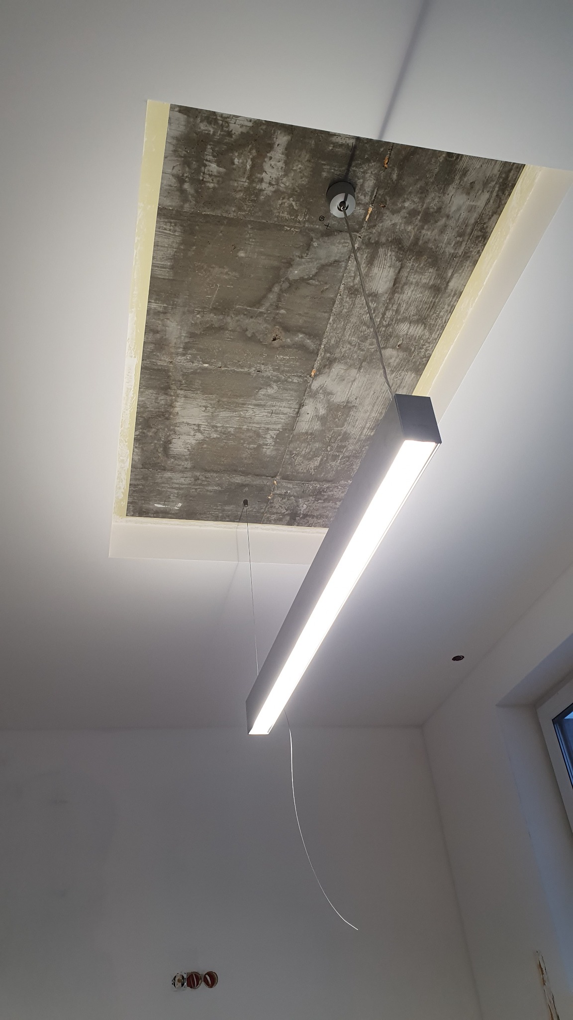 Zawieszona, długa lampa LED w minimalistycznym stylu oświetla surowy, betonowy fragment sufitu z widocznymi śladami szalunku w Pszczółkach, otoczony świeżo pomalowaną, białą powierzchnią.