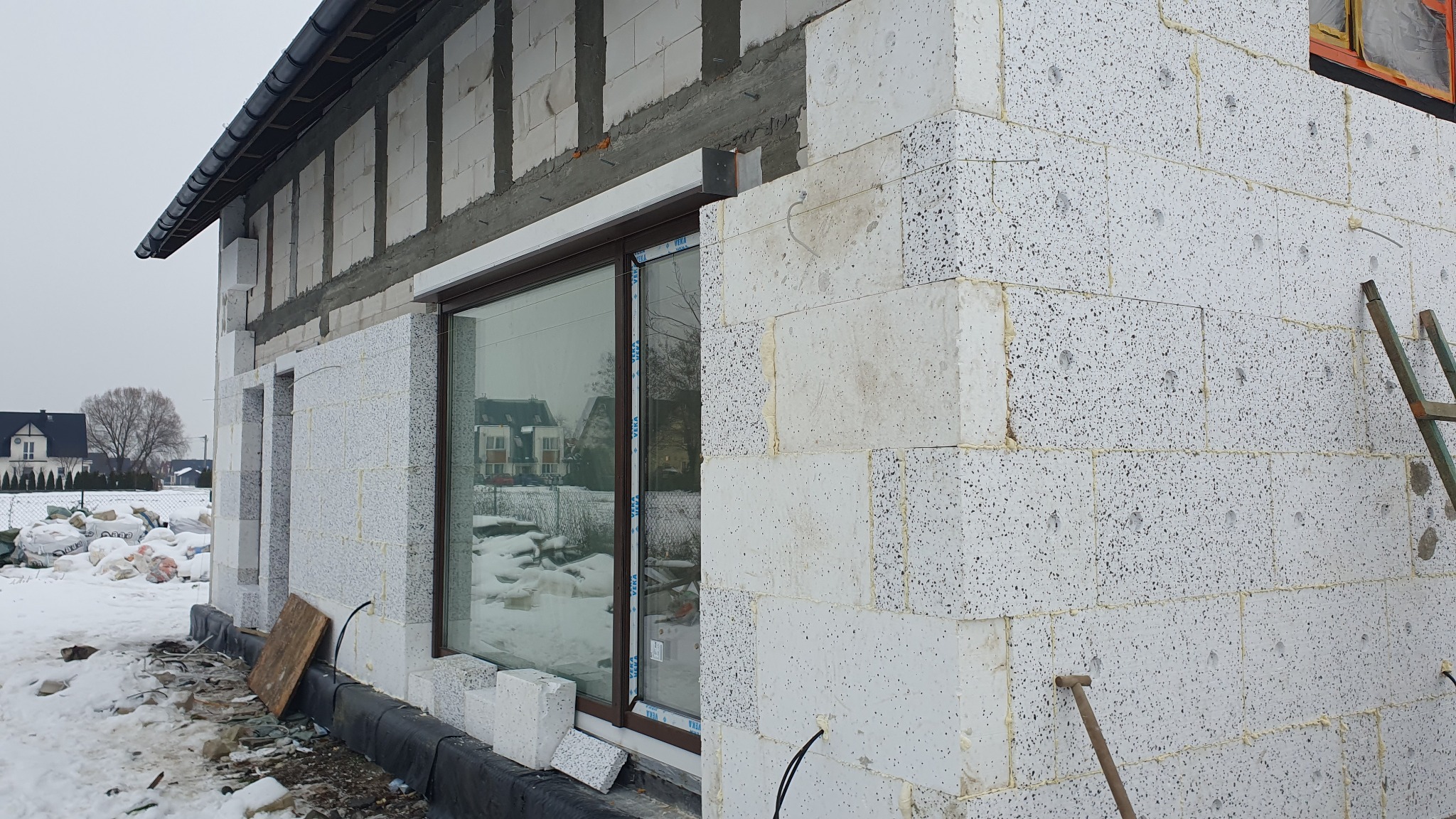 Budowa domu jednorodzinnego w zimowej scenerii, ściany wzniesione z białych bloczków, widoczny otwór okienny z brązową ramą, na zewnątrz śnieg i częściowo ocieplony budynek w tle.