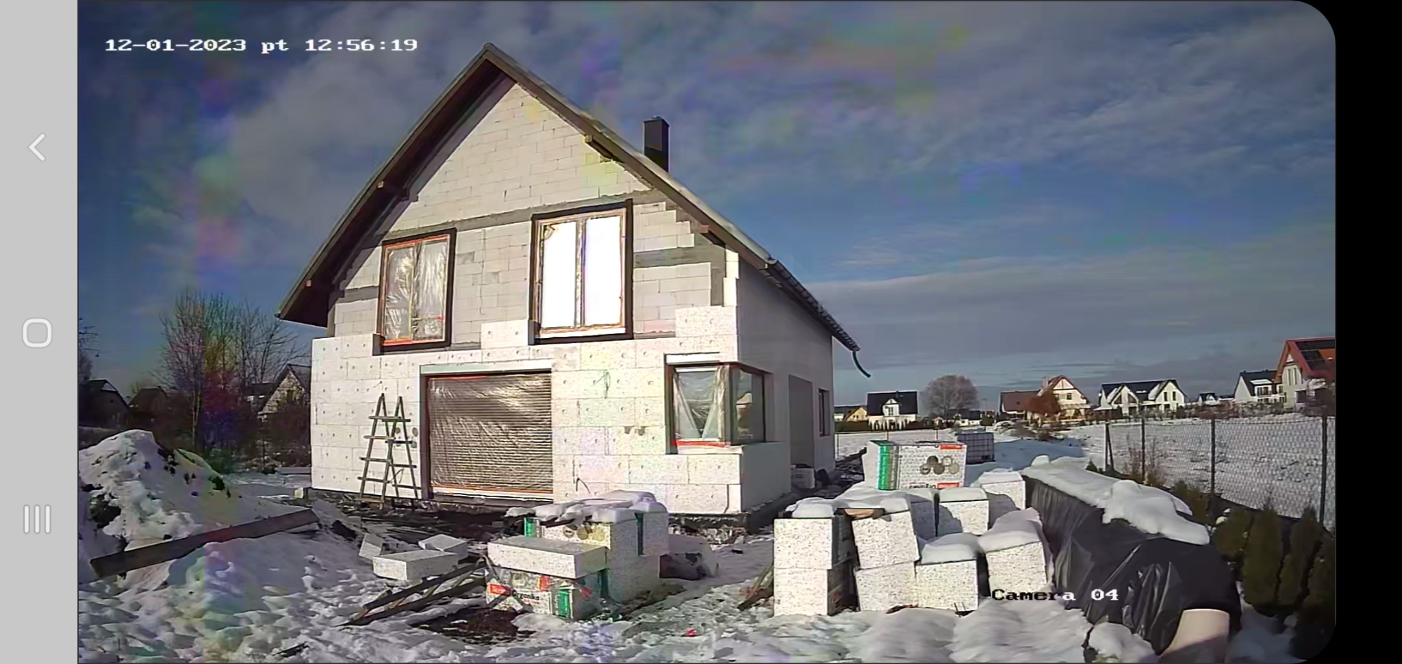 Dom w trakcie ocieplania styropianem, widoczny fragment elewacji z oknami zabezpieczonymi folią, drabina oparta o ścianę, palety styropianu w śniegu, ujęcie z kamery monitoringu.