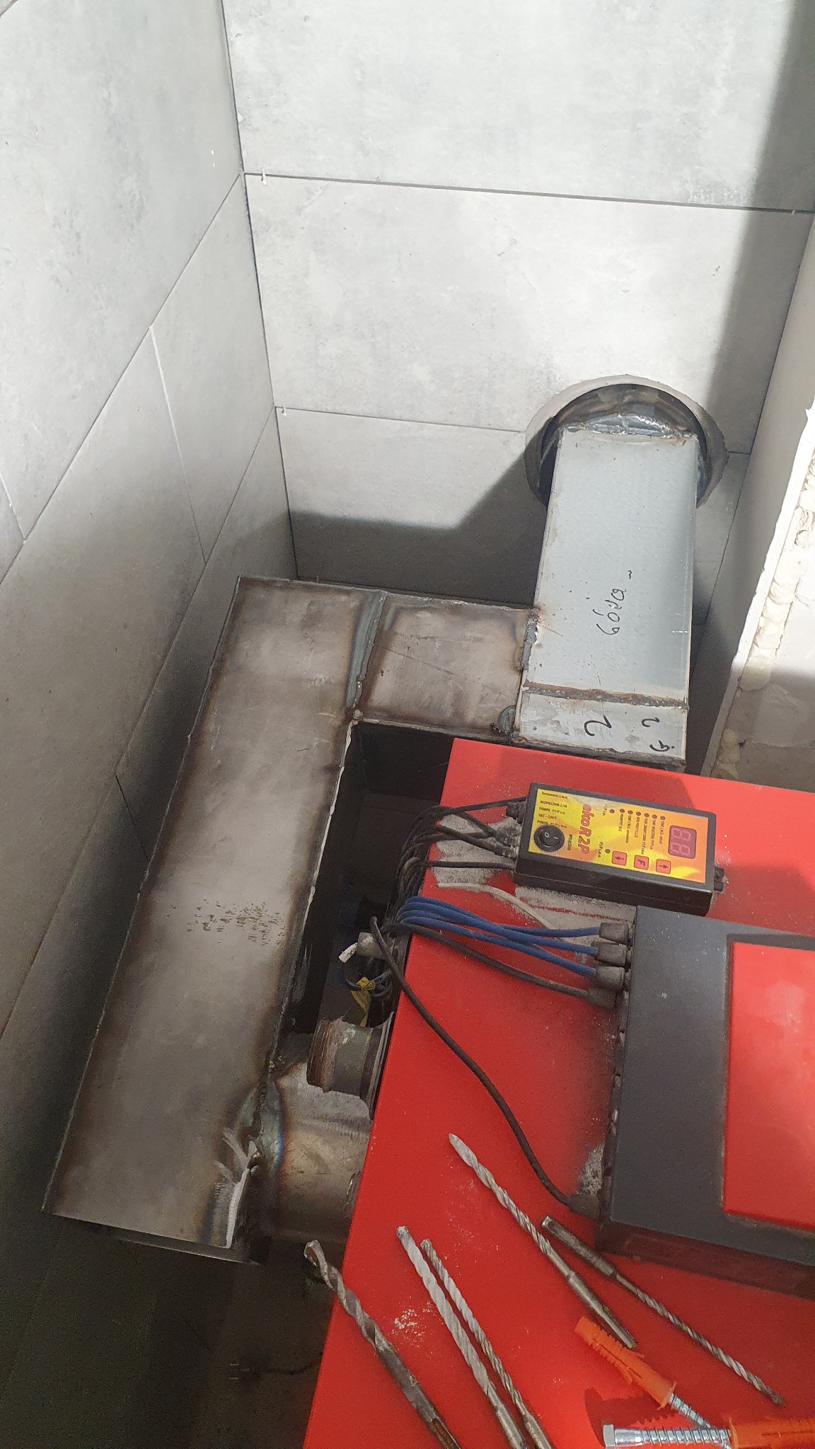 Fragment łazienki z szarymi płytkami, widoczny metalowy komin i czerwony piec z panelami sterowania, obok leżą wiertła i kołki rozporowe.