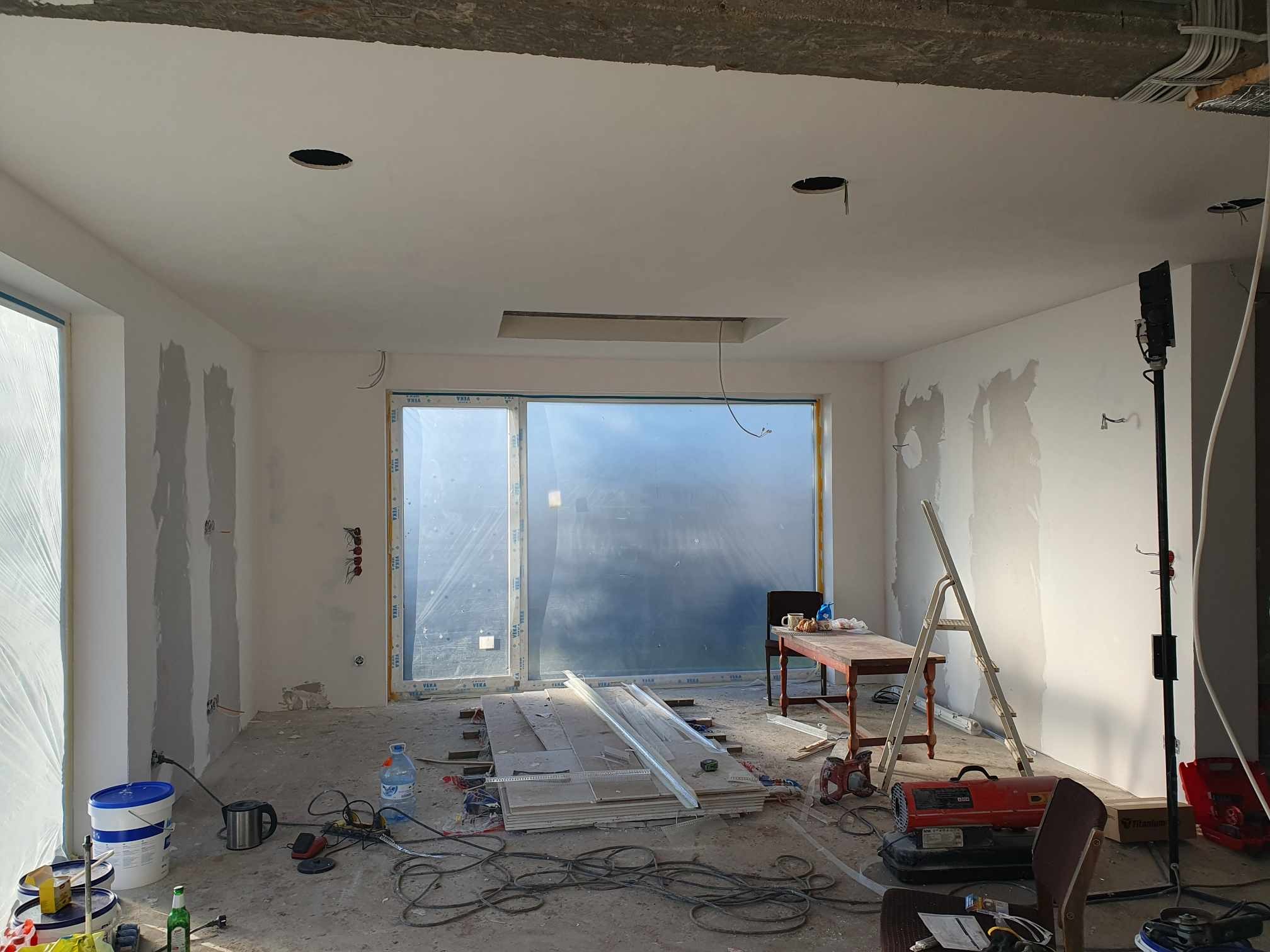 Wnętrze w trakcie prac wykończeniowych: surowe ściany z widocznymi śladami szpachlowania, otwory w suficie pod oświetlenie, rozłożone panele podłogowe, narzędzia i kable na podłodze, drabina oparta...