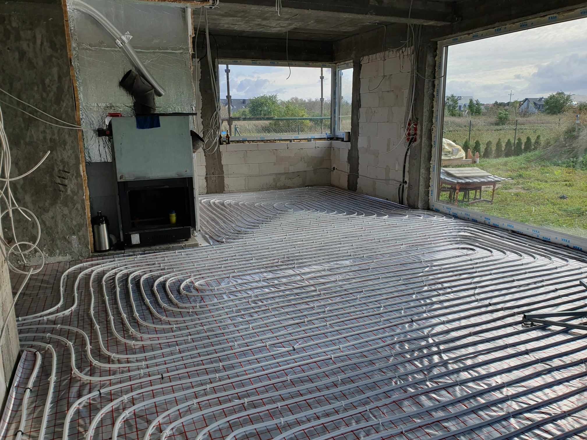 Surowy pokój z widocznym systemem ogrzewania podłogowego rozłożonym na folii izolacyjnej, kominek w stanie surowym, widok przez okno na zewnątrz.