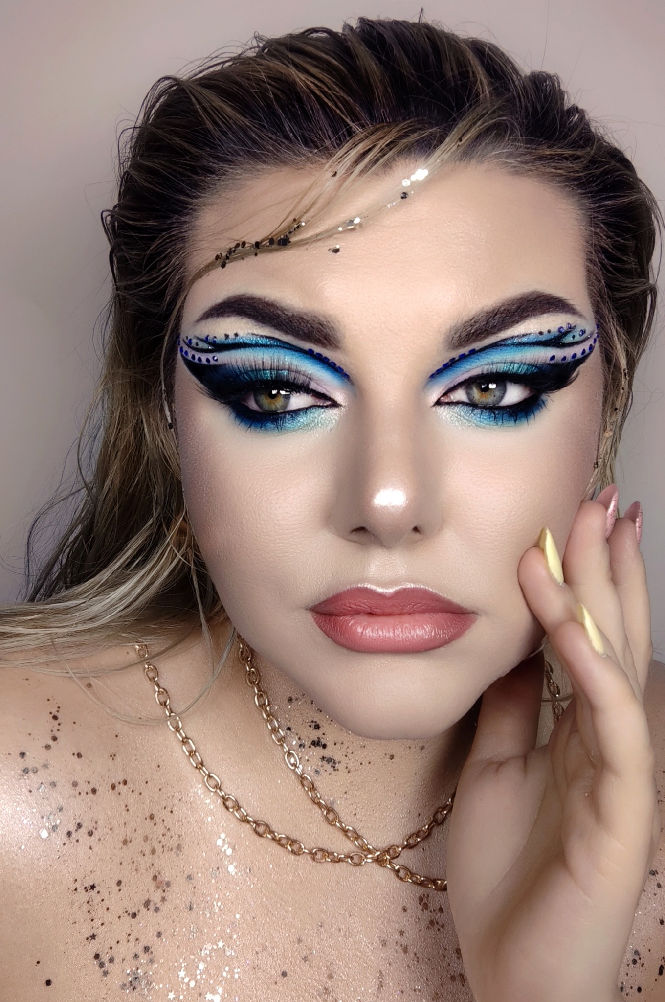 Portret kobiety z intensywnym, niebiesko-czarnym zdobieniem powiek, brokatem na ciele i włosach, oraz złotym łańcuszkiem na szyi.