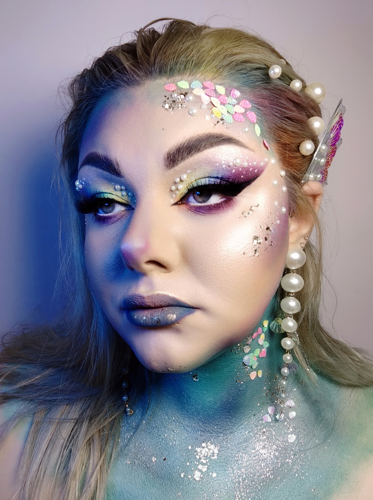 Portret kobiety z artystyczną stylizacją inspirowaną syreną: niebiesko-zielony body painting, perłowe ozdoby na włosach i twarzy, brokat oraz pastelowe muszelki na czole i szyi, intensywny kolorowy...