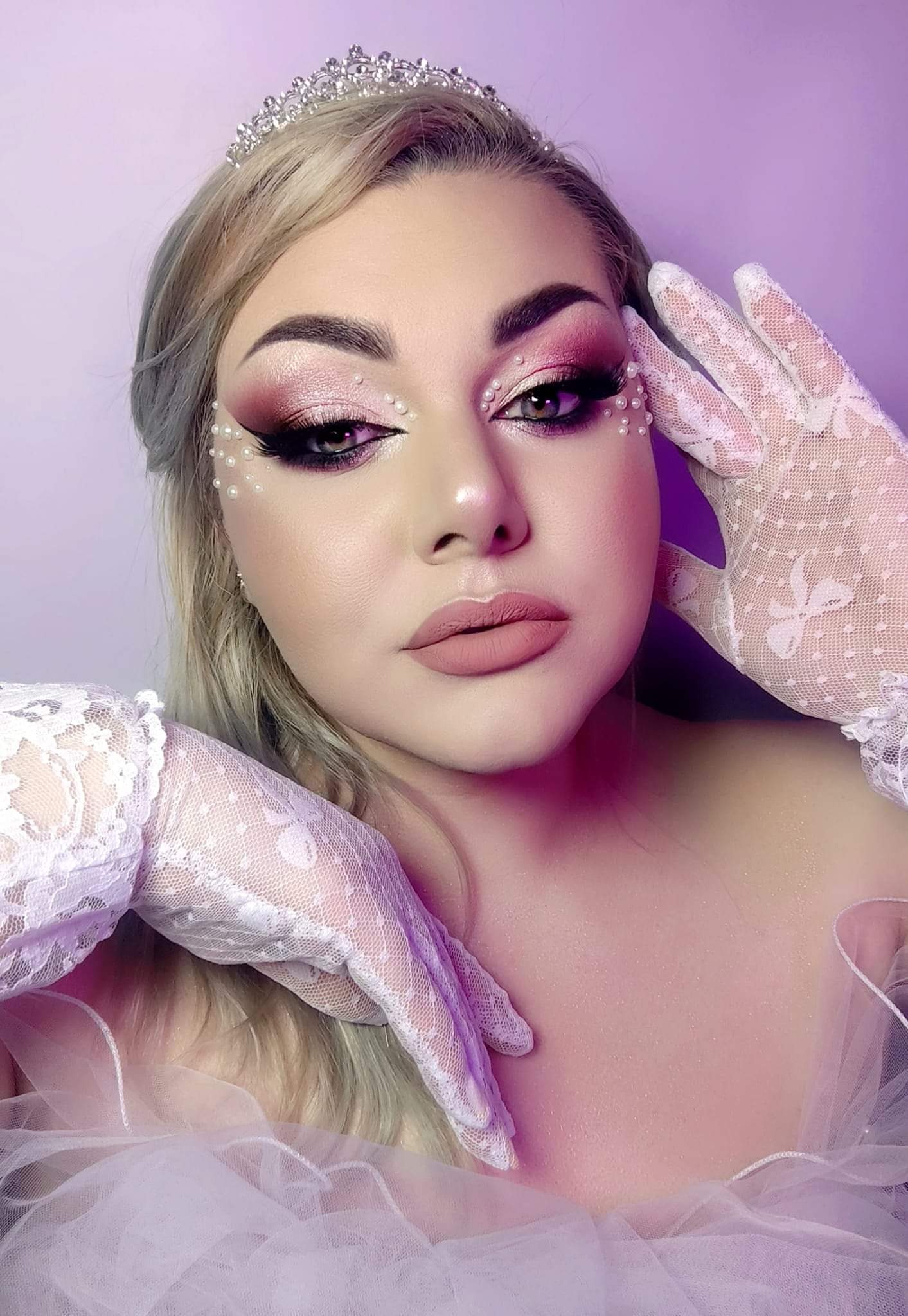 Portret kobiety z delikatnym, różowym cieniowaniem powiek, sztucznymi rzęsami, perłami wokół oczu i ust, w tiulowej sukni i koronkowych rękawiczkach, z tiarą na głowie, na fioletowym tle.