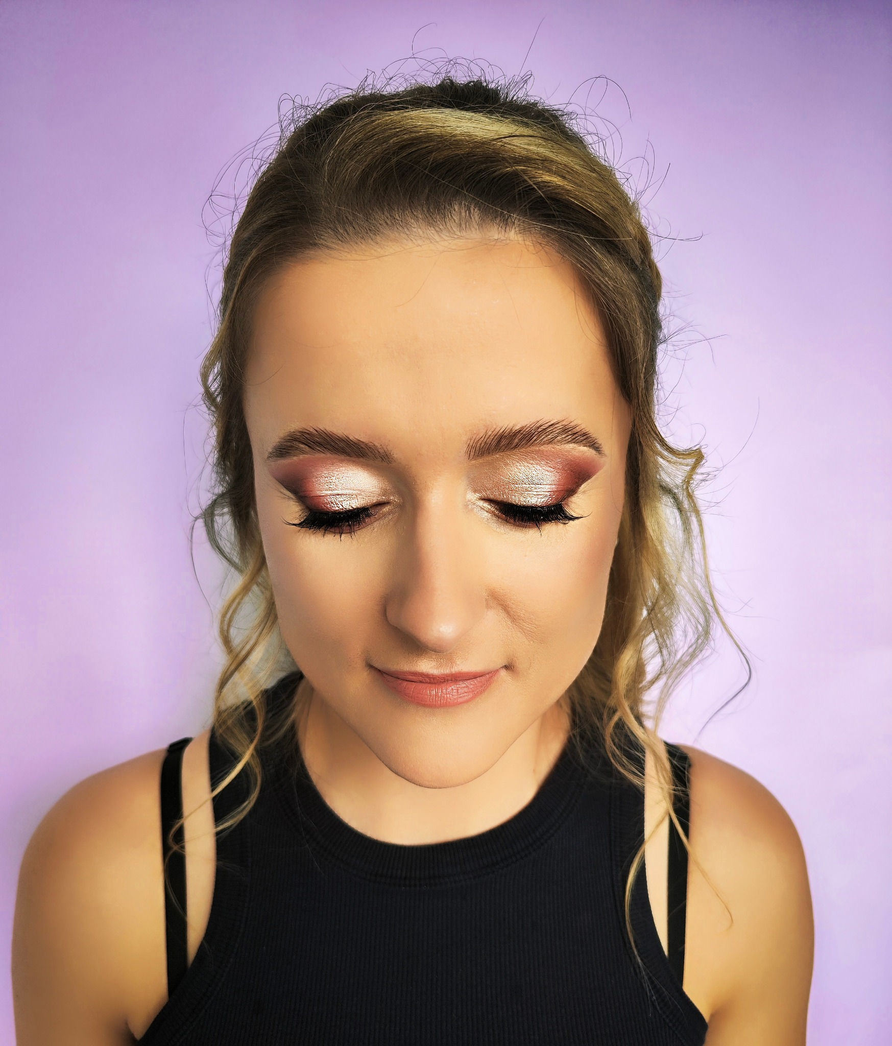 Portret kobiety z zamkniętymi oczami, prezentujący błyszczący, różowo-złoty cień do powiek z wyraźnie podkreślonymi rzęsami na fioletowym tle.