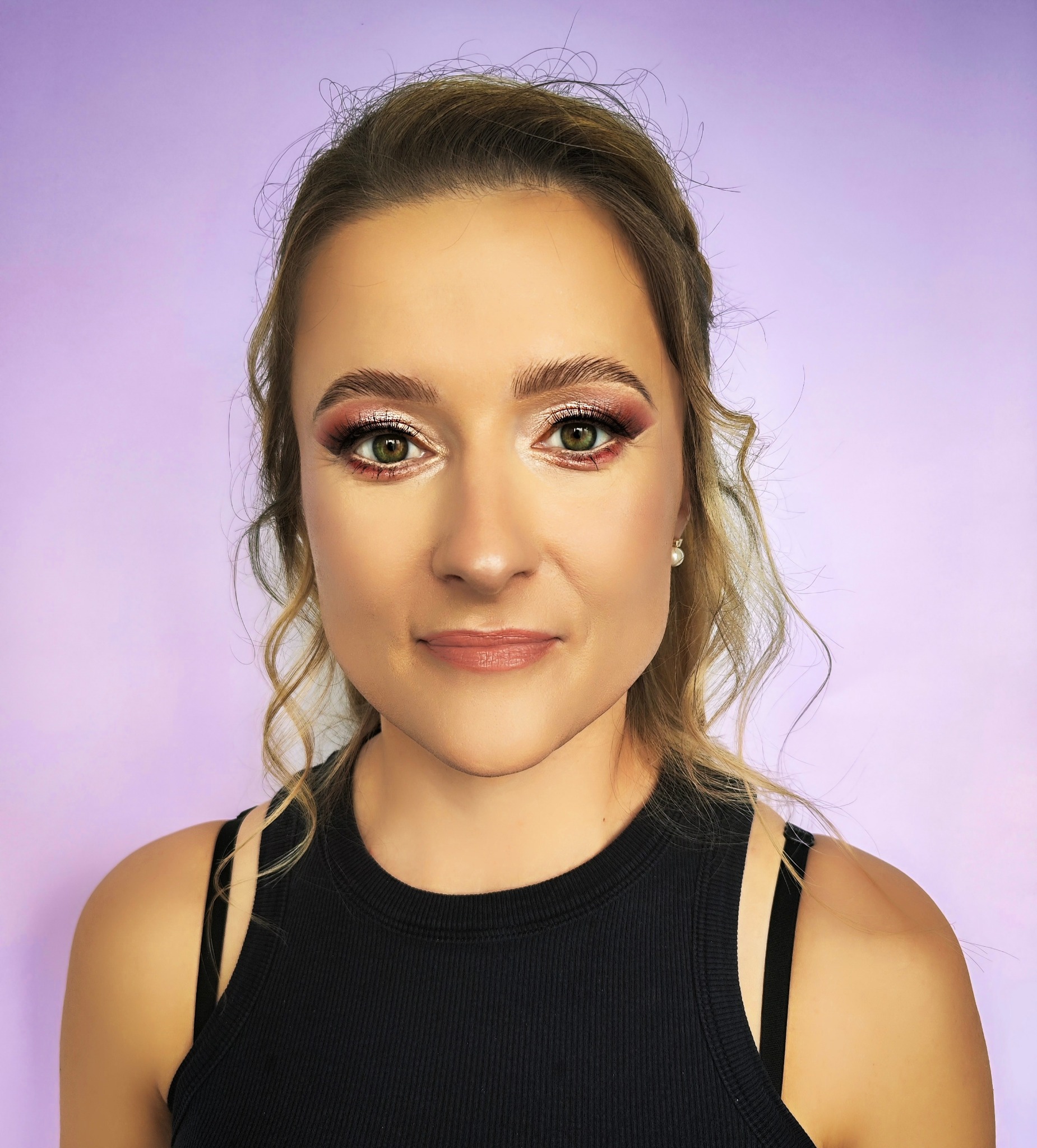Portret kobiety z profesjonalnym makijażem oczu w odcieniach różu i złota, delikatnie podkreślone usta, z zielonymi oczami i blond włosami upiętymi z boku, na fioletowym tle.