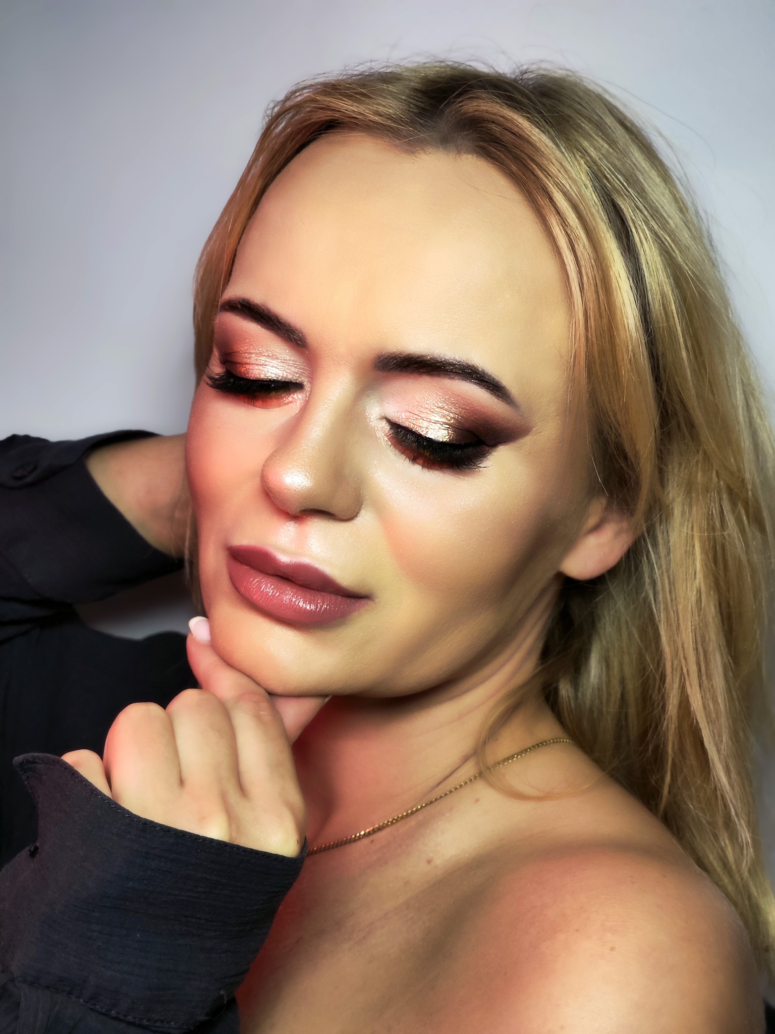 Portret kobiety z zamkniętymi oczami, prezentującej profesjonalnie wykonany, błyszczący, brązowo-złoty odcień na powiekach oraz podkreślone usta w odcieniu nude z połyskiem. Delikatna biżuteria...